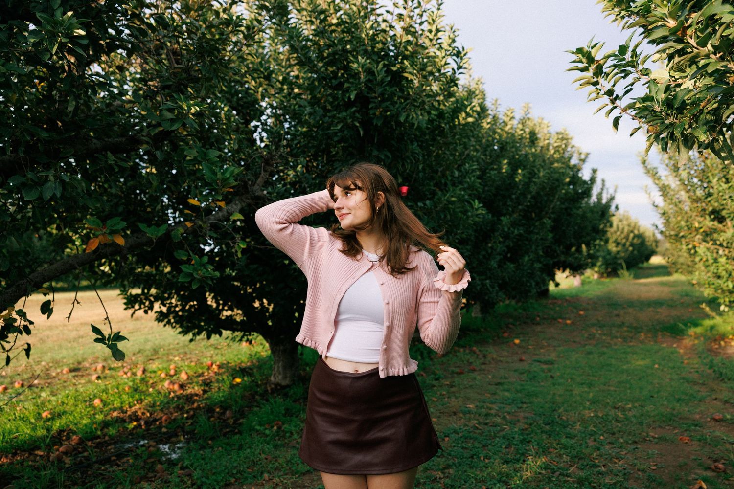 Someone poses in a pink jacket and brown skirt among fruit trees in an orchard.