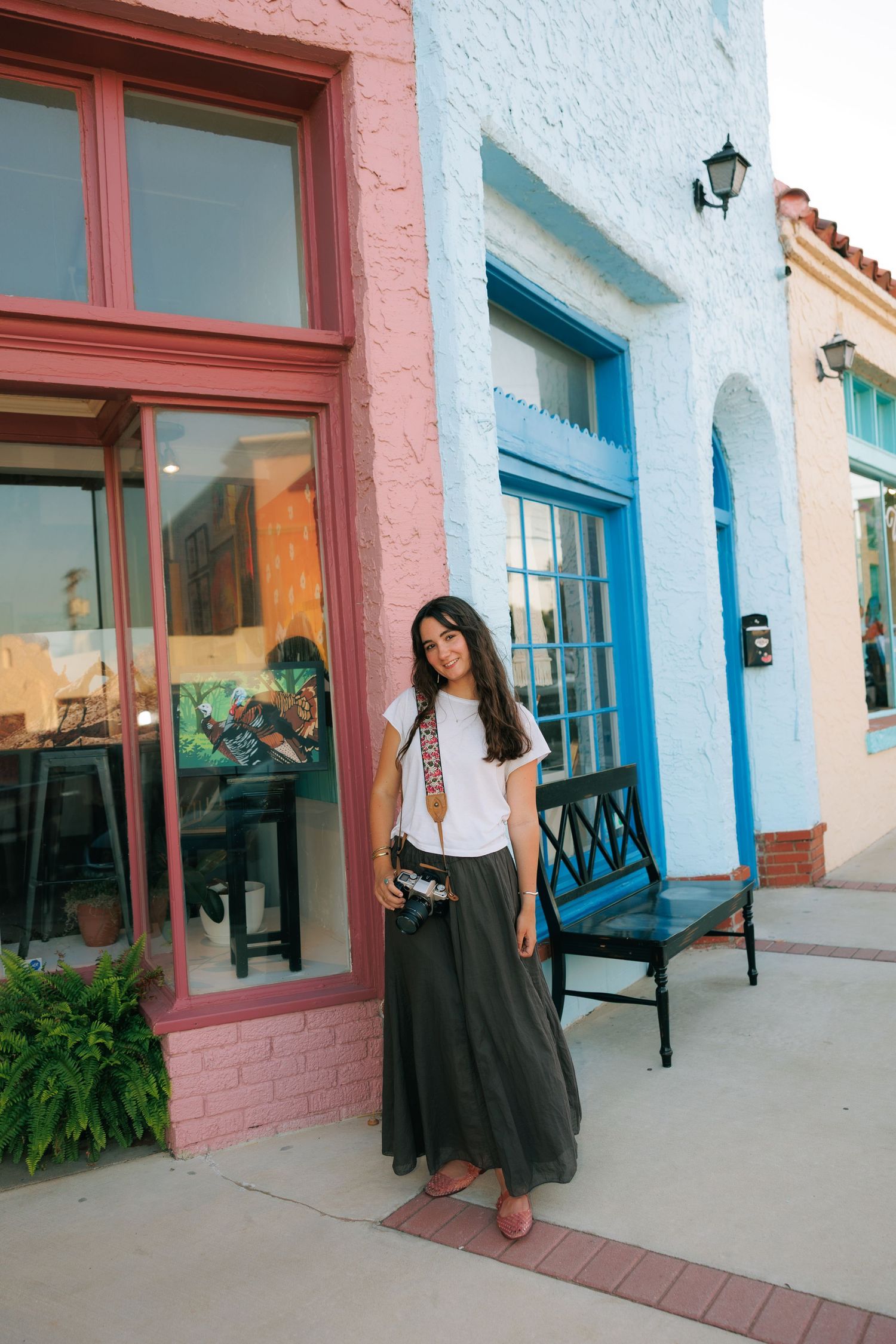 Woman in white tee and olive maxi skirt posing outside colorful pink and blue storefronts.