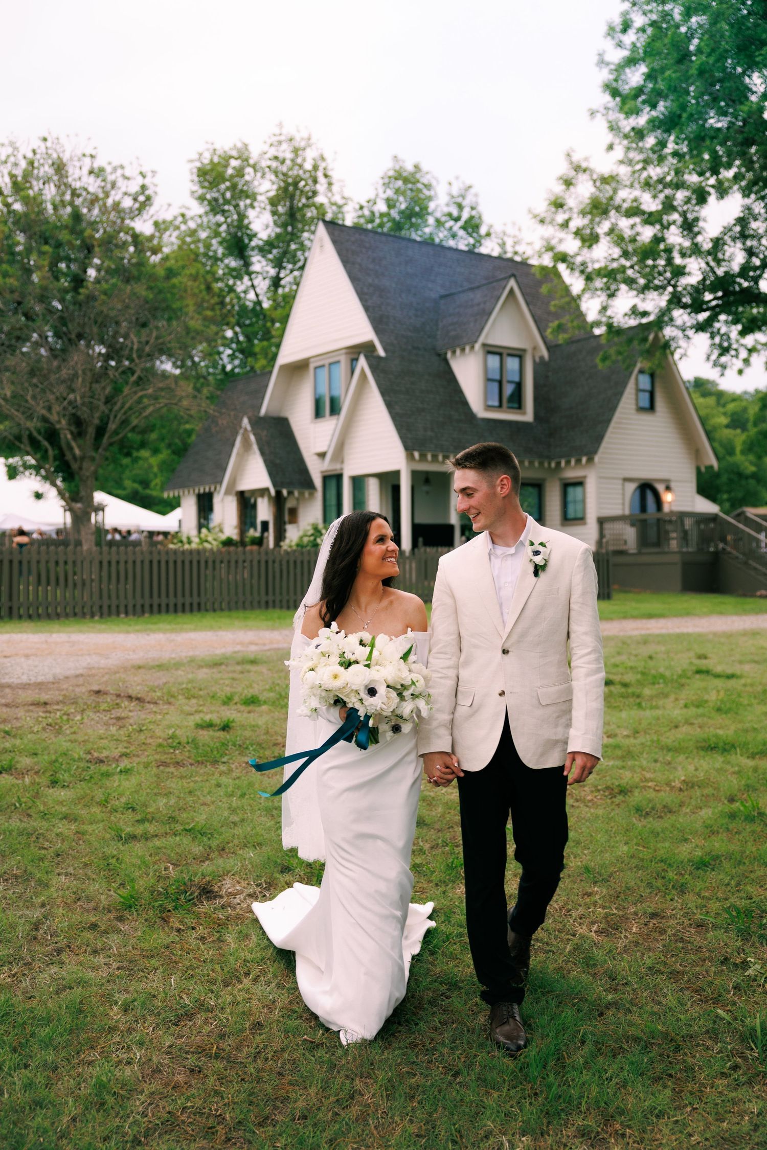 Couple in white formal wear walking together on grassy lawn with farmhouse in background.