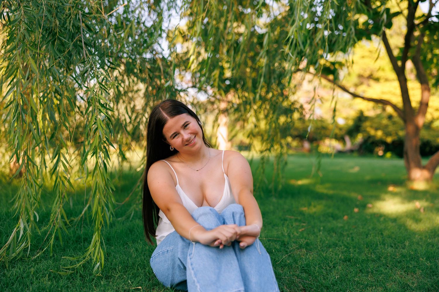 Person in blue dress sitting on grass under willow tree branches in summer garden.