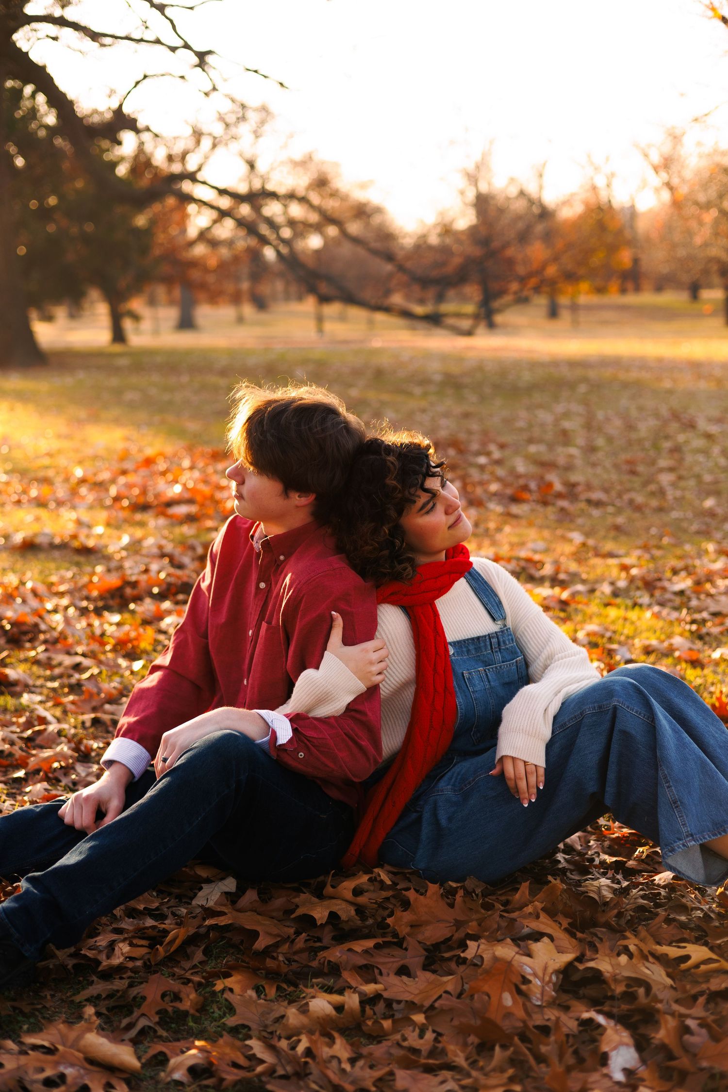 Two people in red and blue outfits relax on fallen autumn leaves during golden hour sunset.