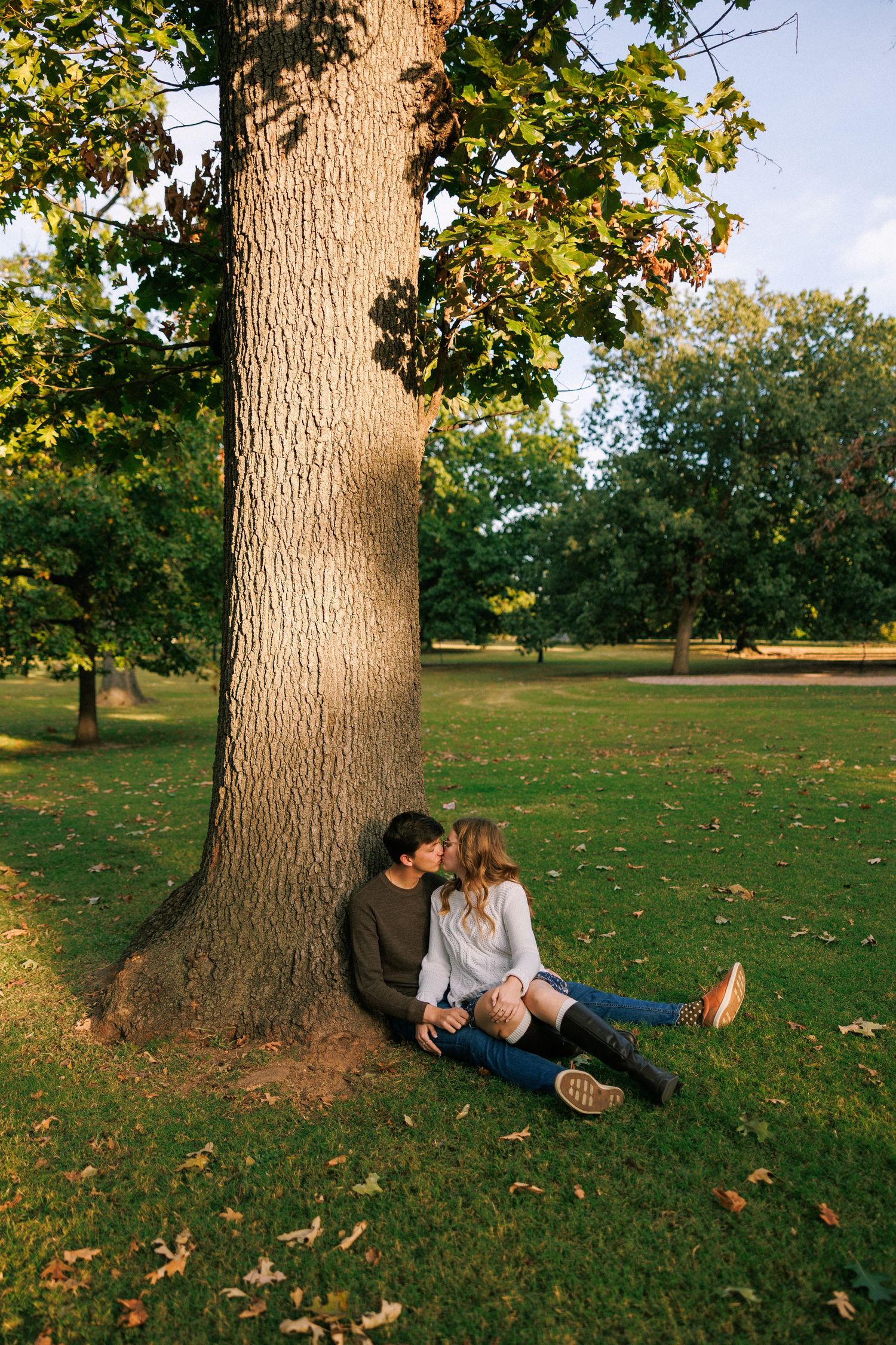 A couple sits together at the base of a large tree trunk in a serene park setting during autumn.