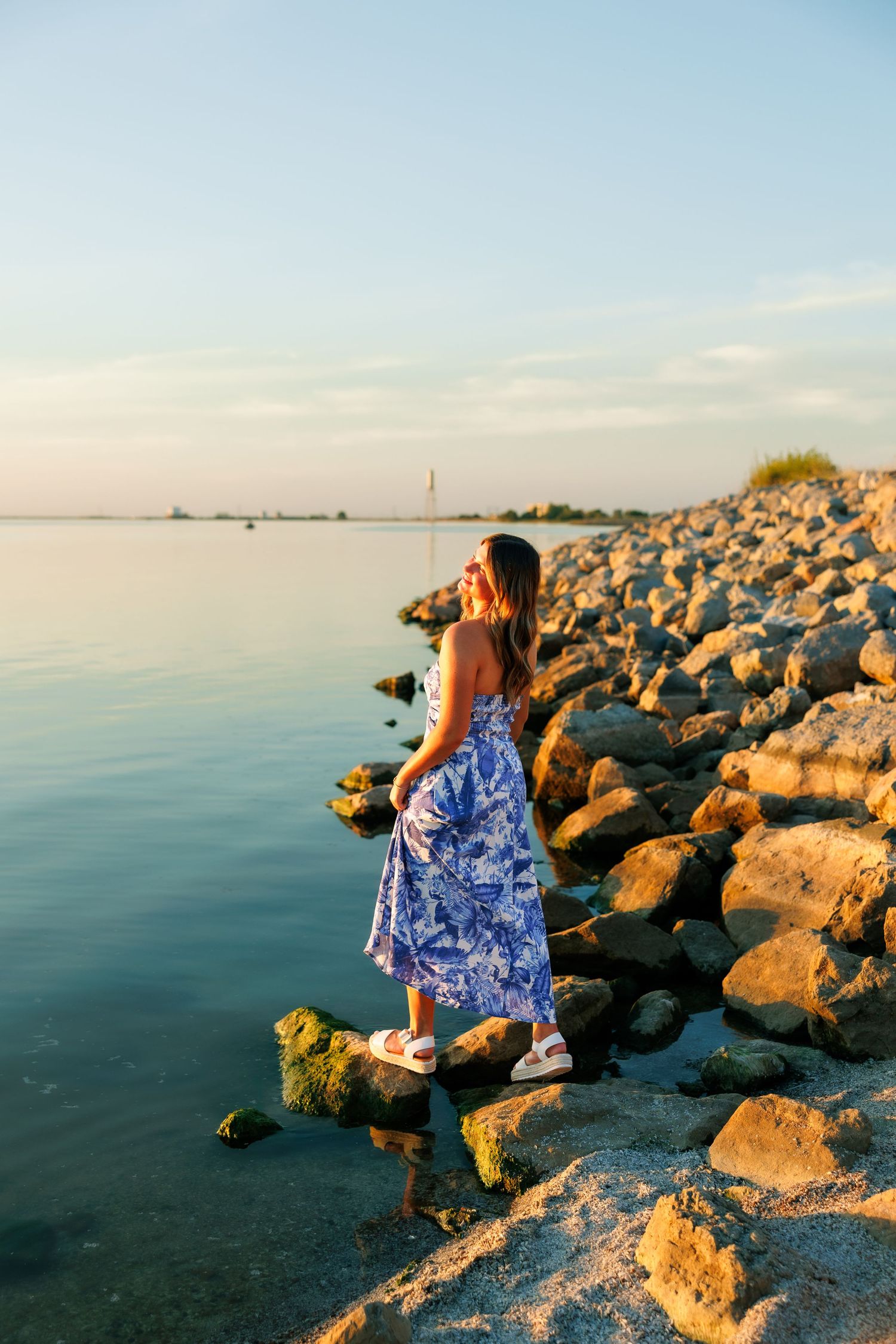 A flowing blue dress catches the breeze along a rocky shoreline at golden hour.