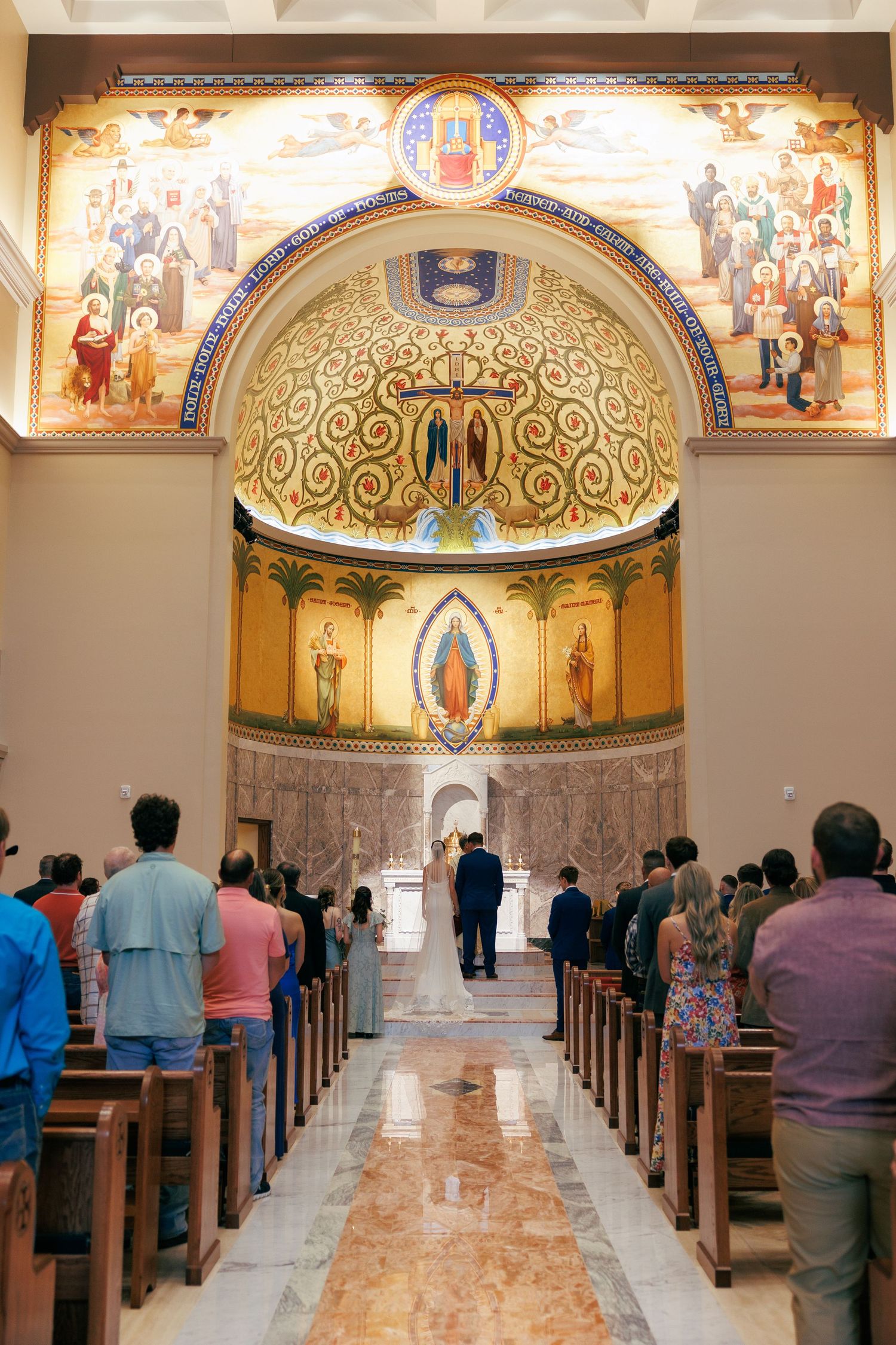 Interior of an ornate Catholic church with marble floors and decorated dome ceiling during a wedding ceremony.