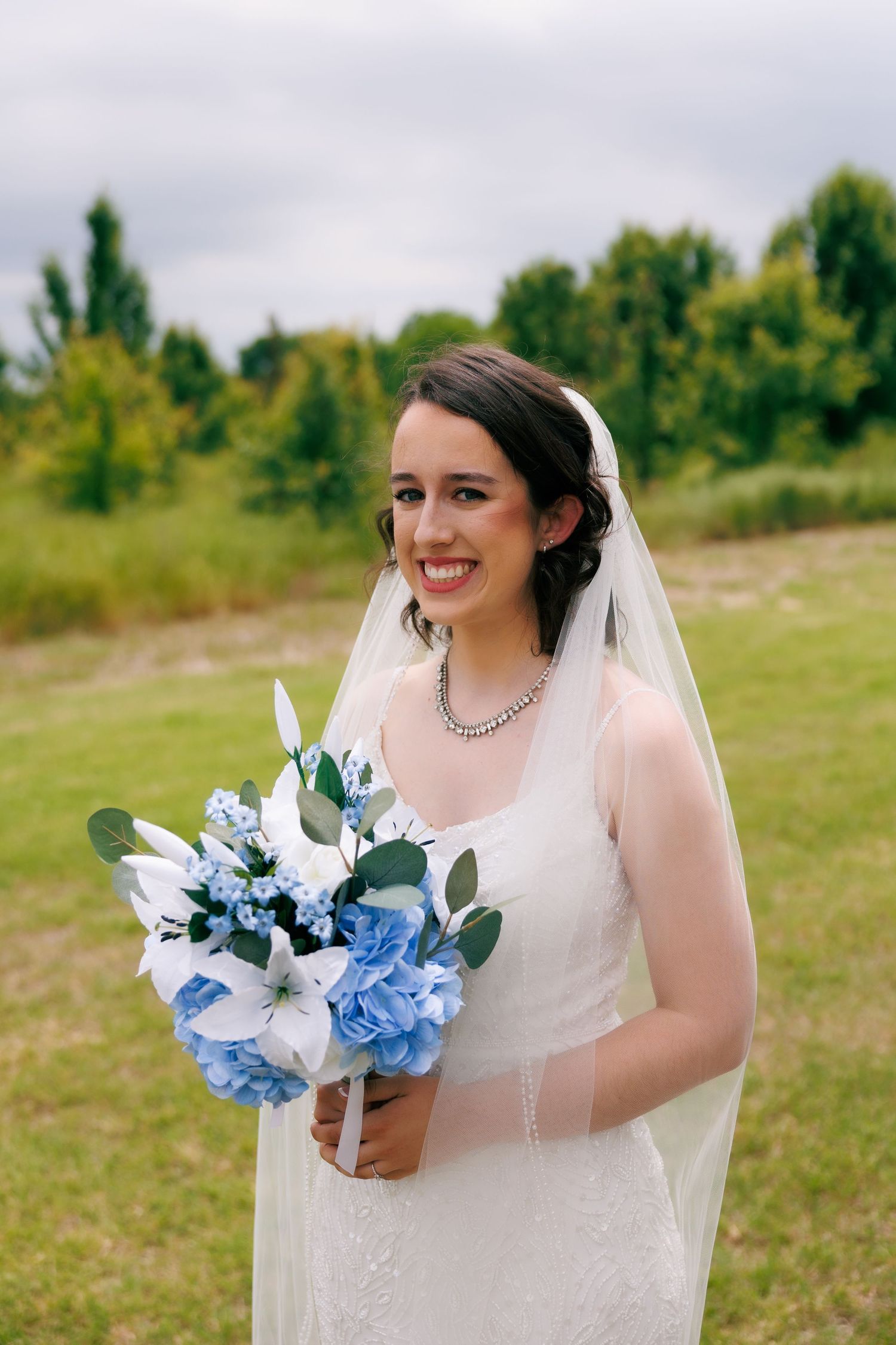 A bride in white dress holds a blue and white bouquet in a sunny outdoor field.