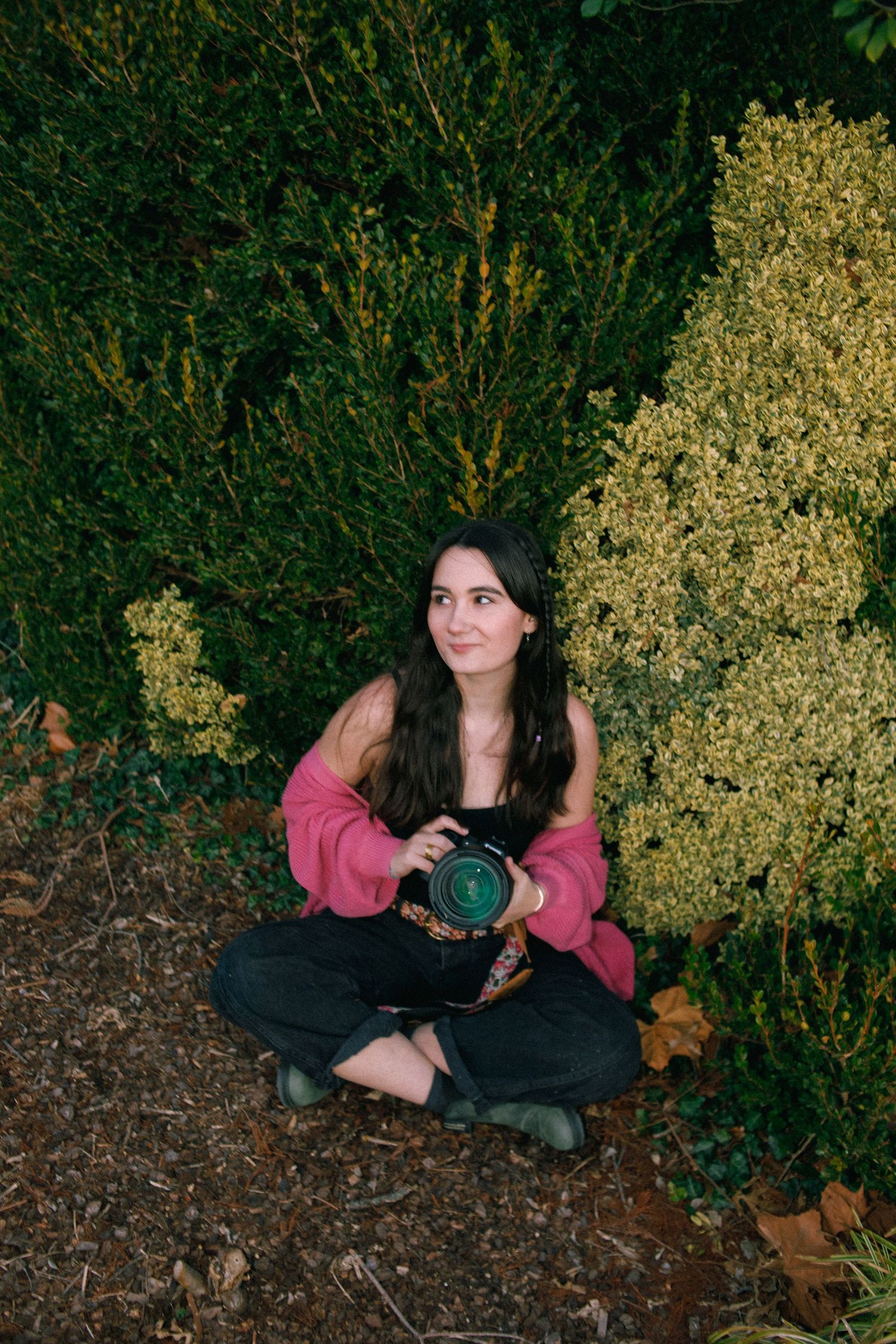 A photographer in a pink sweater sits cross-legged on the ground with camera equipment near flowering bushes.