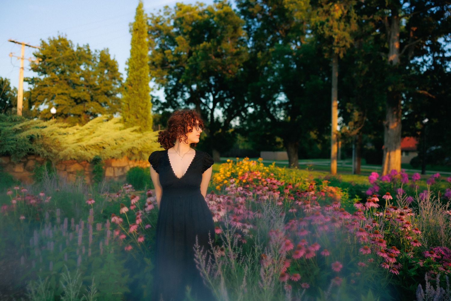 Woman in black dress standing among colorful blooming flowers in a lush green garden at golden hour.