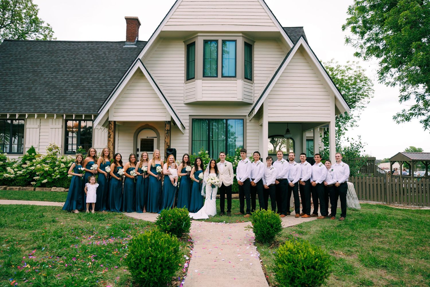 Large group portrait of wedding party gathered in front of white chapel-style building.