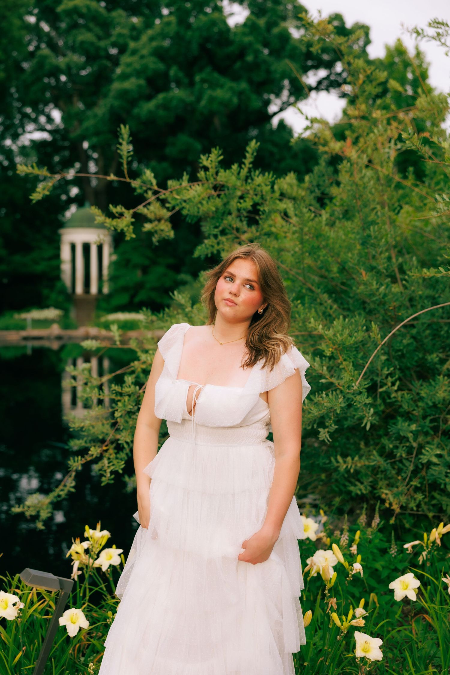 Young woman in white tulle dress standing among yellow lilies in a lush garden with a pond and gazebo.