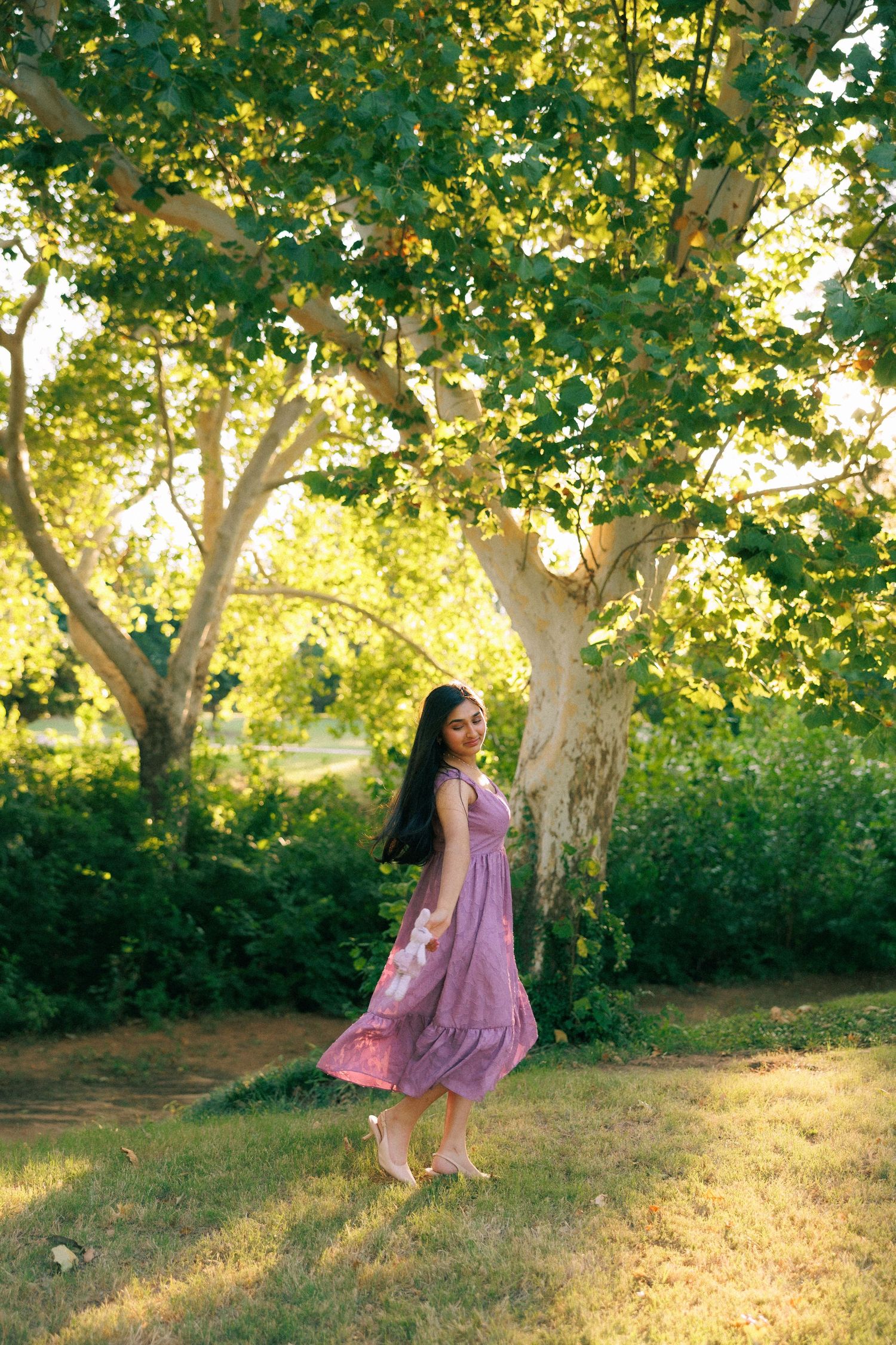 Young woman in purple dress standing barefoot under green trees in golden sunlight in a lush park setting.