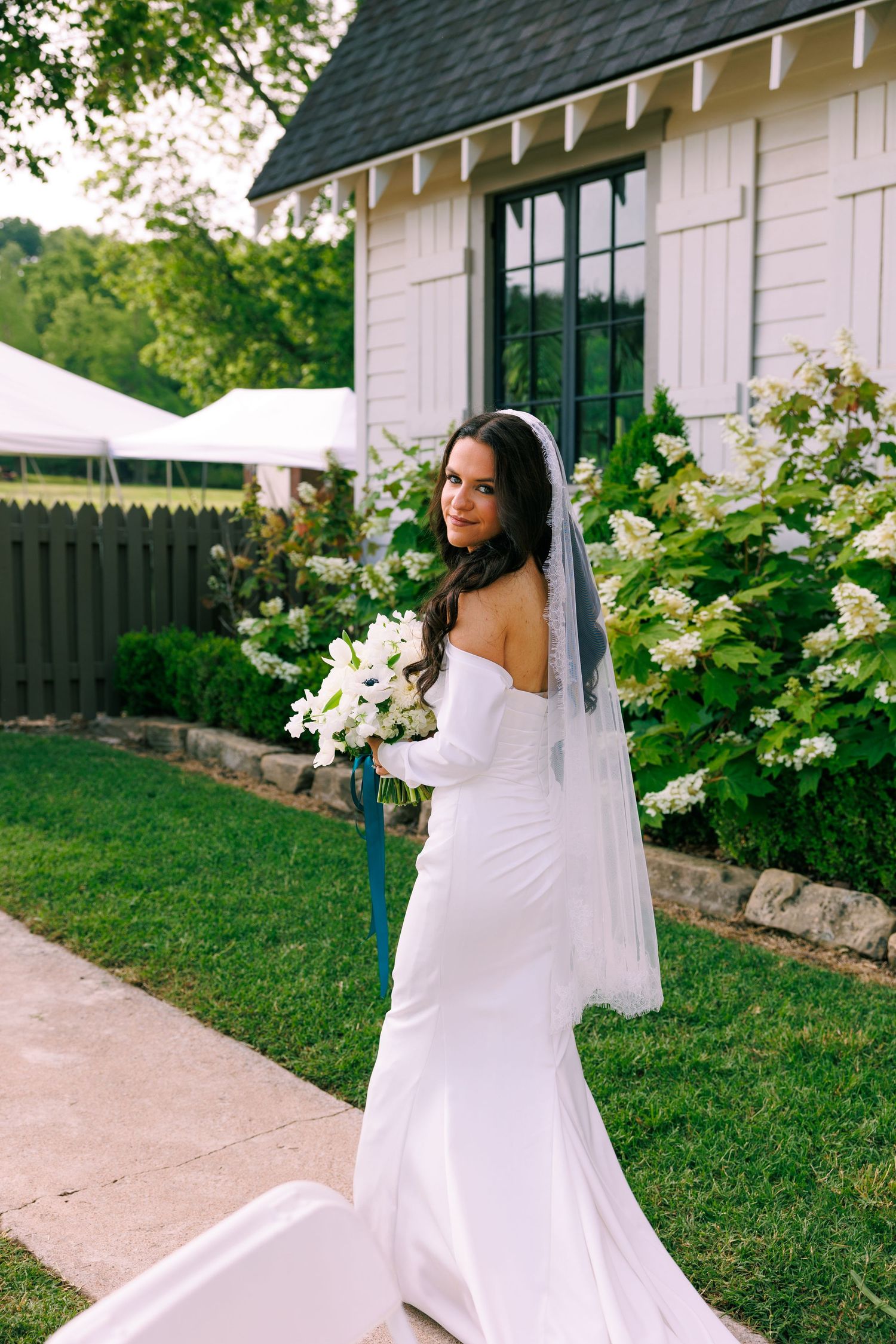 Bride in flowing white dress poses on pathway near white house with green landscaping.