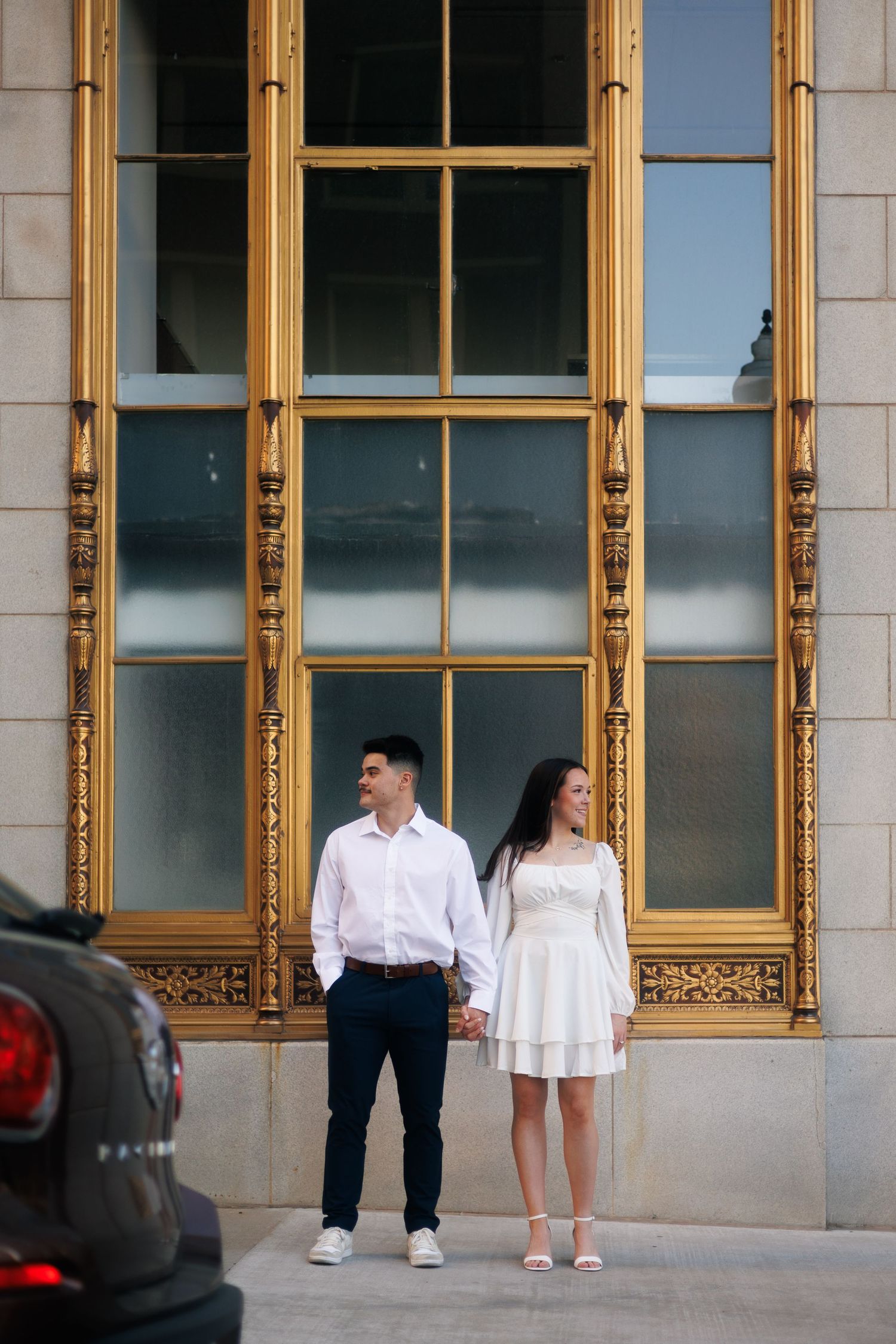 Couple in white attire walking together outside building with golden-framed windows.