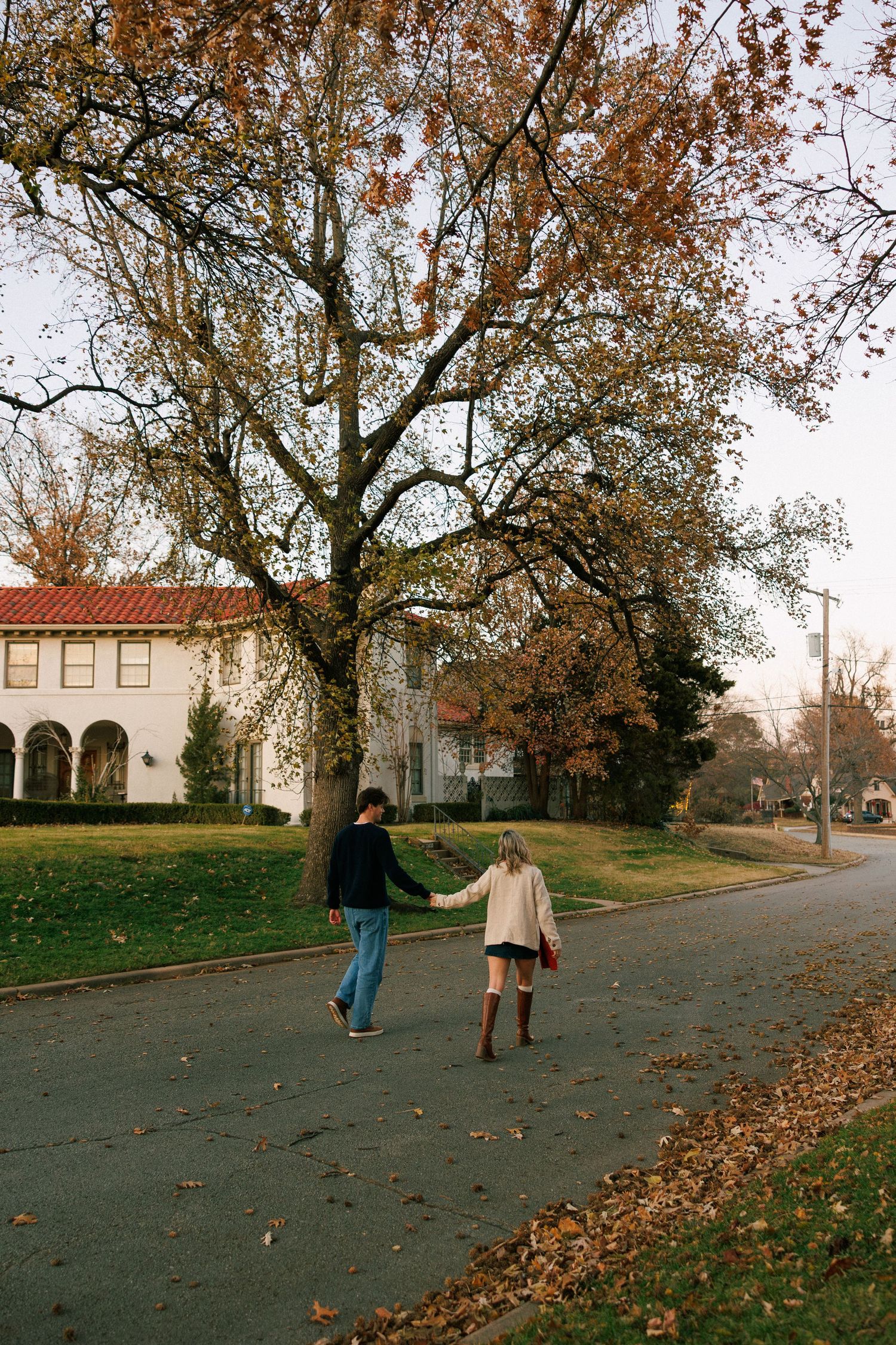 Two people walking along a path near a Mediterranean-style building with autumn trees overhead.