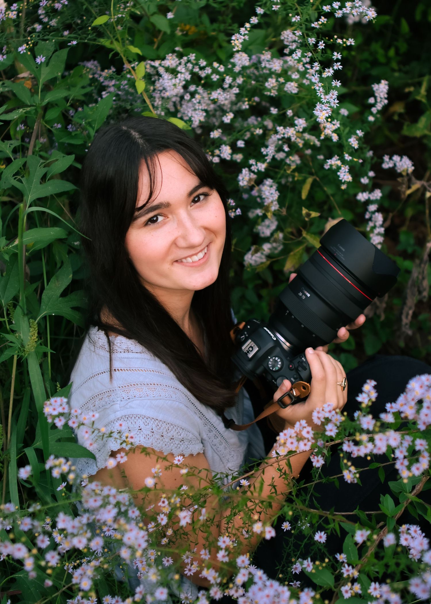 A photographer with a camera poses among delicate purple wildflowers in a garden.