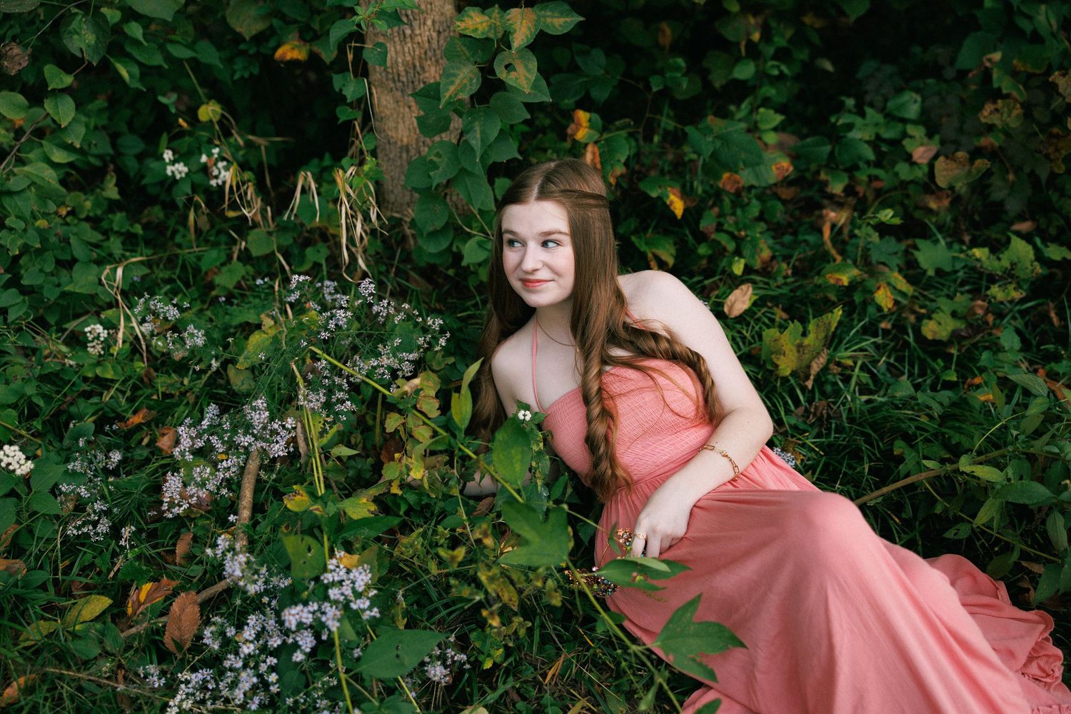 A person in a pink dress poses playfully against green foliage in a garden setting.