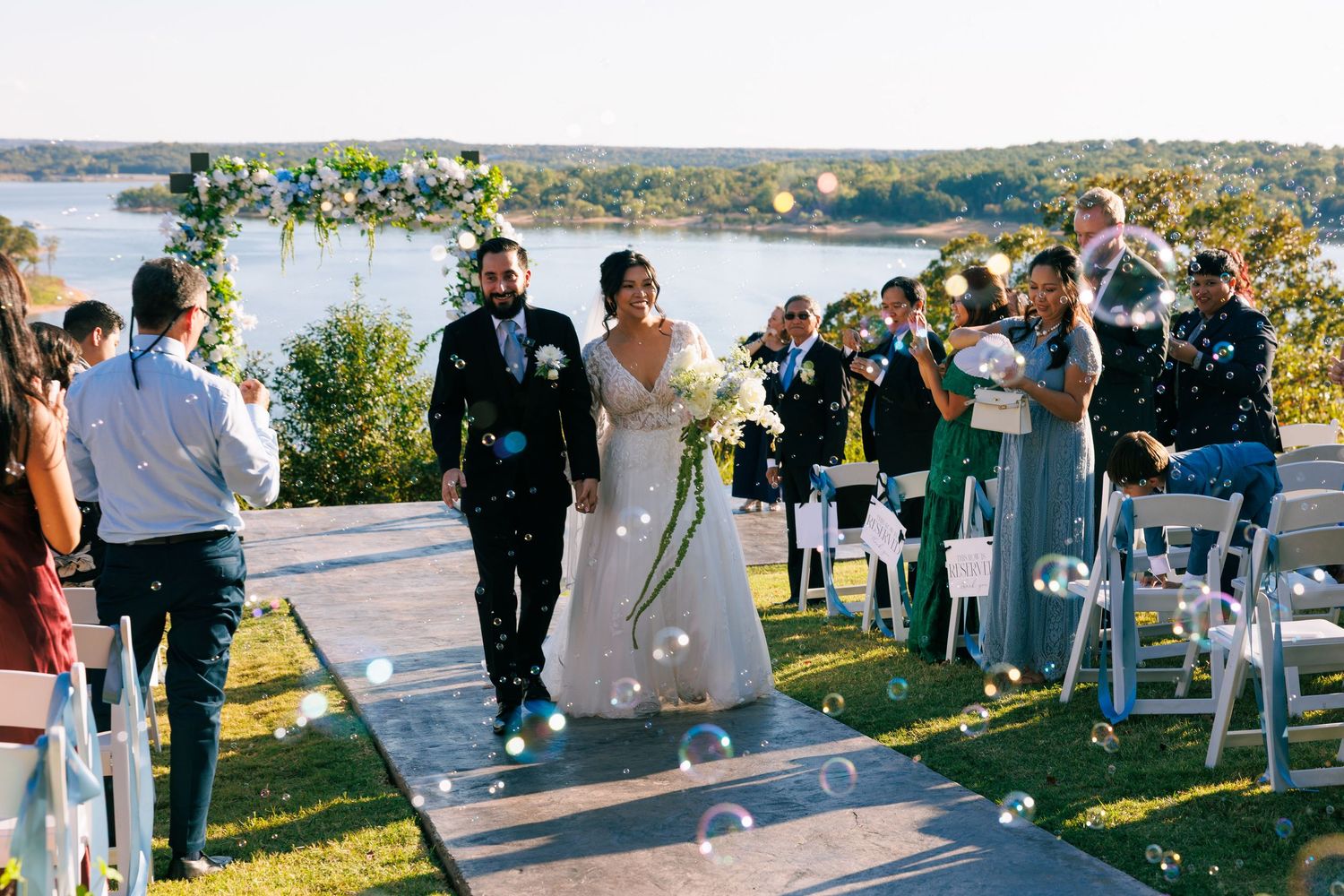 Wedding guests celebrate and throw confetti as the newlyweds walk down the aisle by a lake.