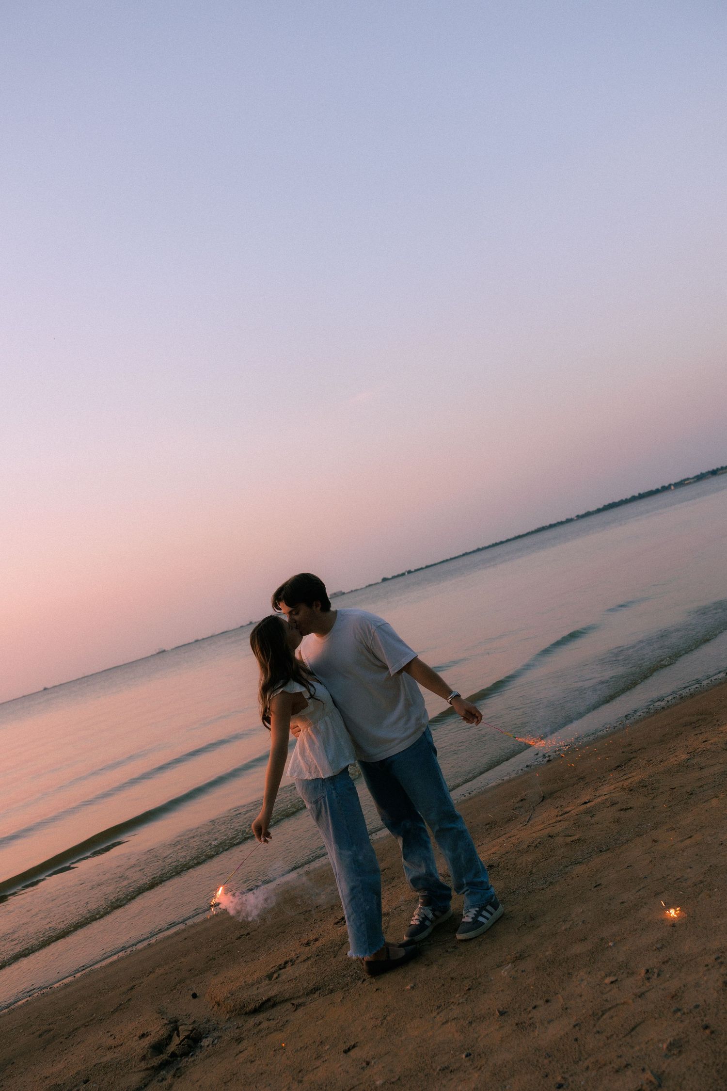 Couple sharing a romantic kiss on a sandy beach at sunset with soft pink and purple hues reflecting on calm water.