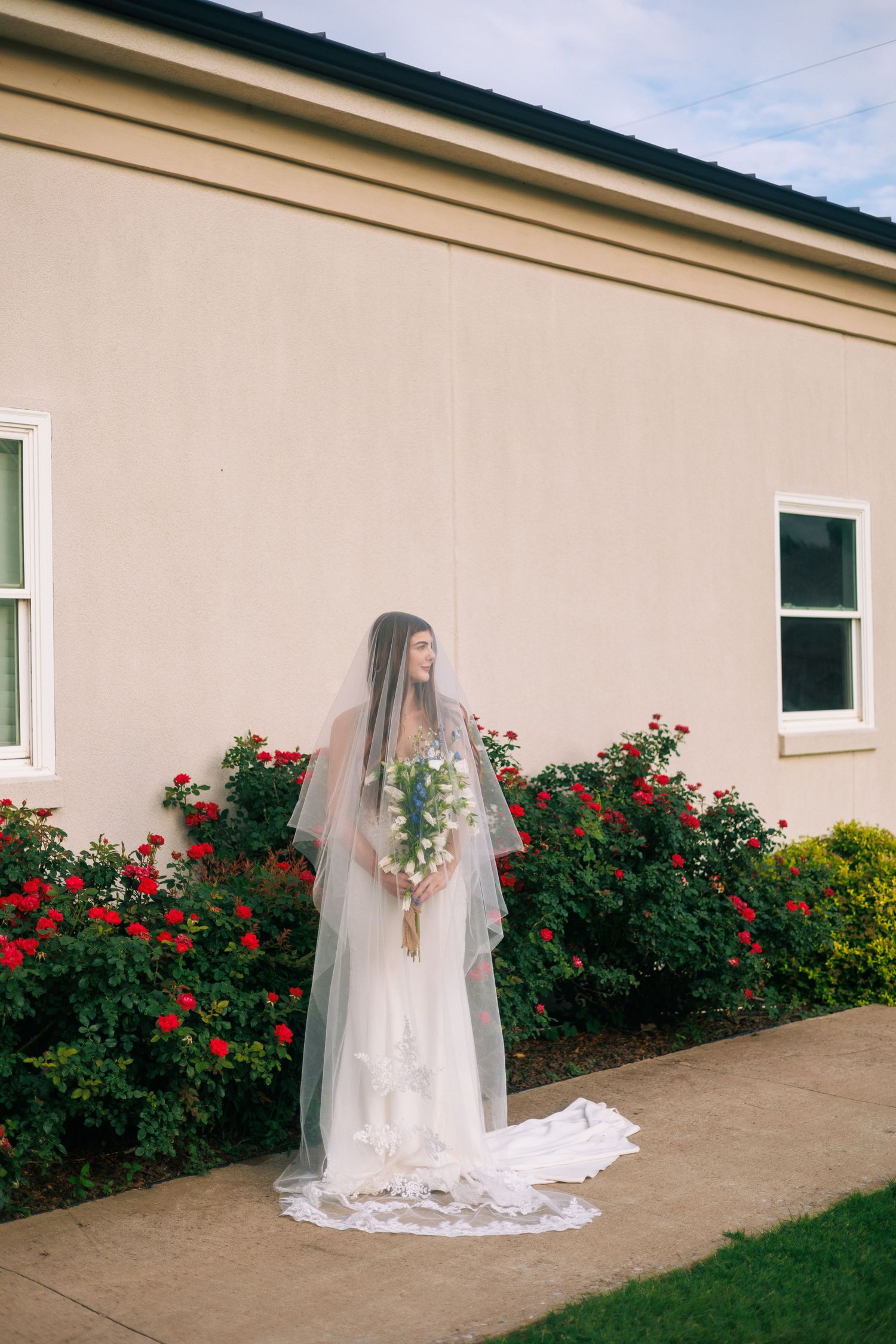 A bride in a flowing white gown poses against a wall with vibrant red roses.