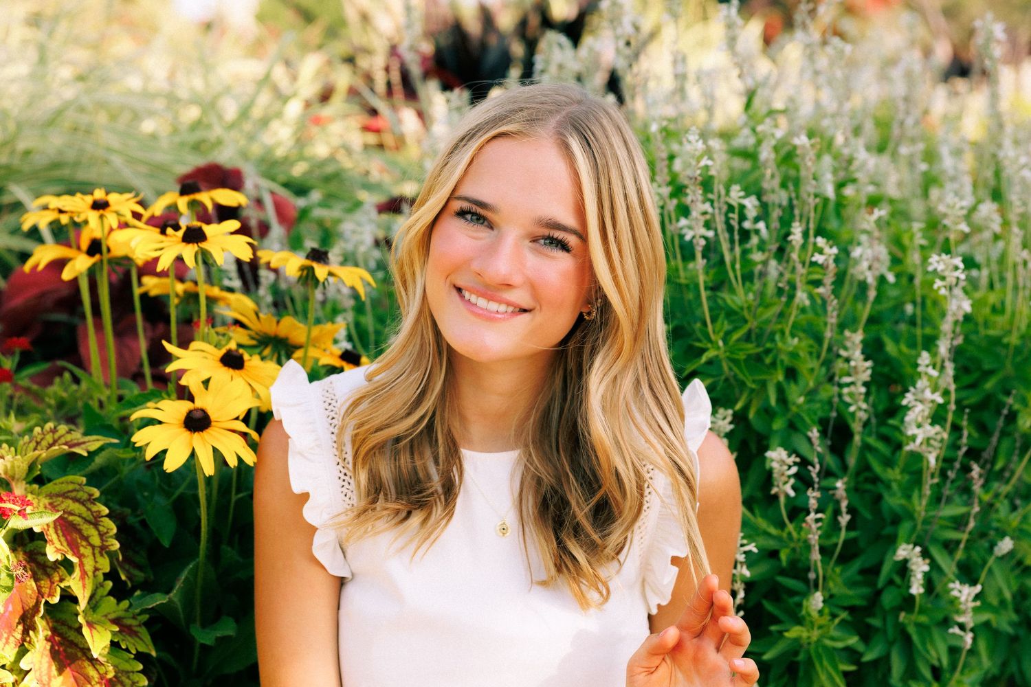 Smiling young woman with blonde hair posing in a vibrant garden with yellow and purple wildflowers.