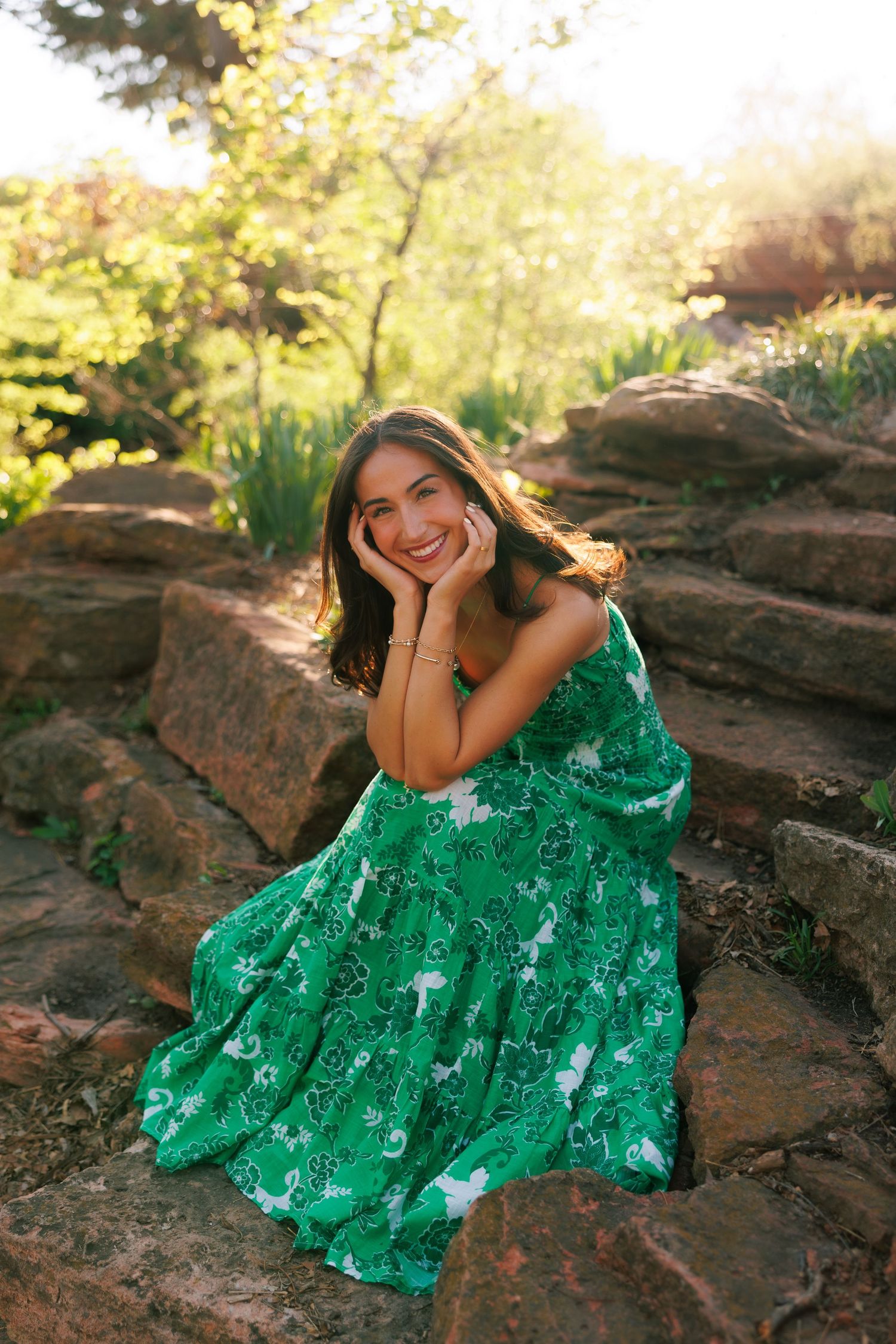 Young woman in green floral dress sitting on stone steps, smiling in a sunlit garden.