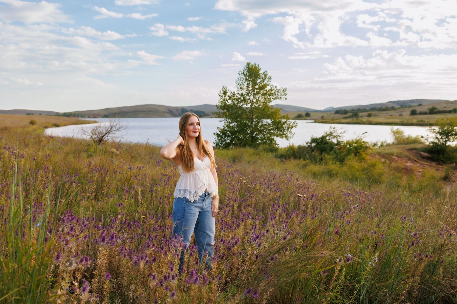 Person in denim and white top stands among purple wildflowers with lake view in background.
