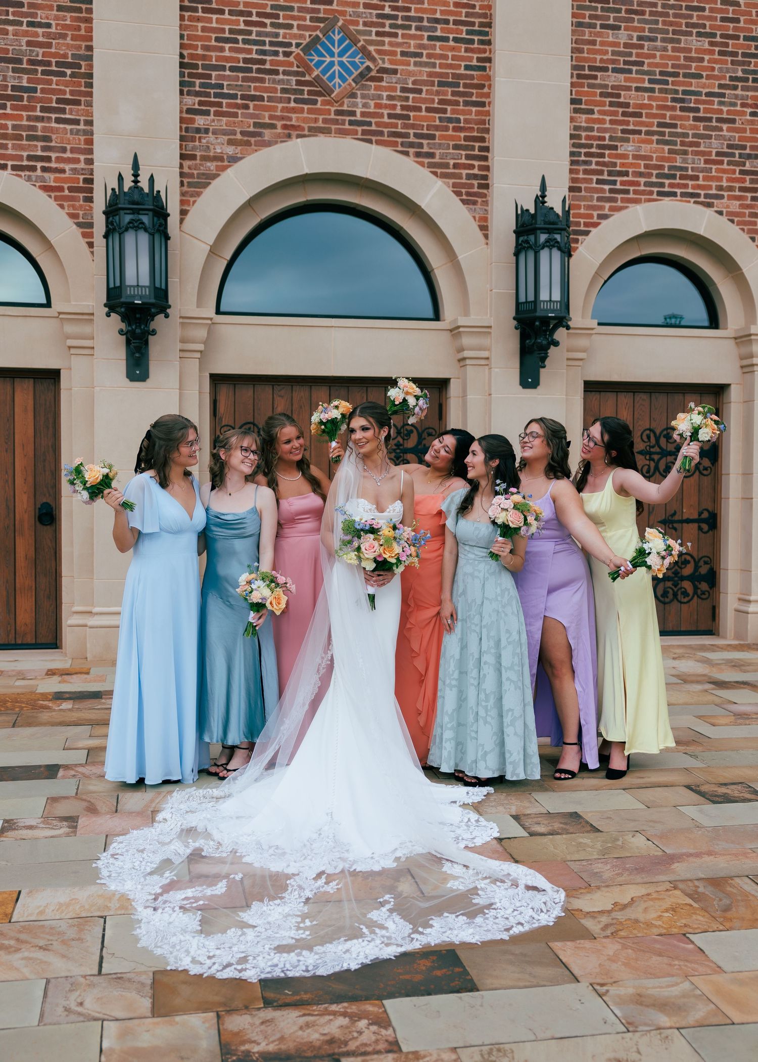 Bridesmaids in pastel dresses pose with bride against brick building with arched windows.