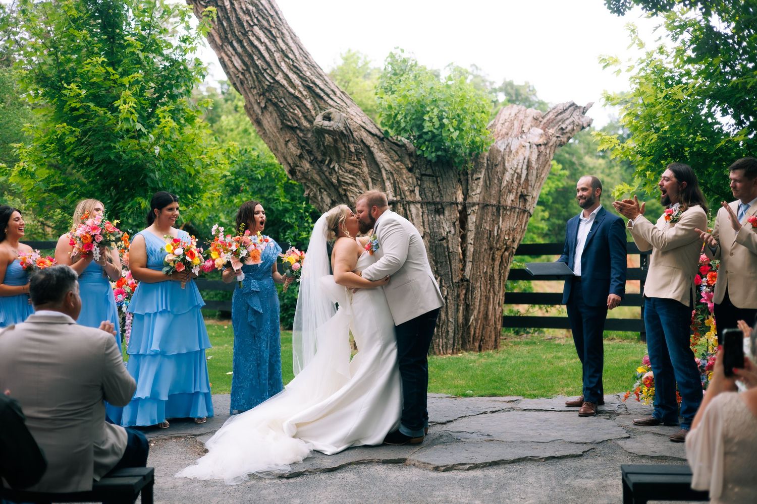 Wedding ceremony takes place under a large tree with guests seated on wooden chairs.