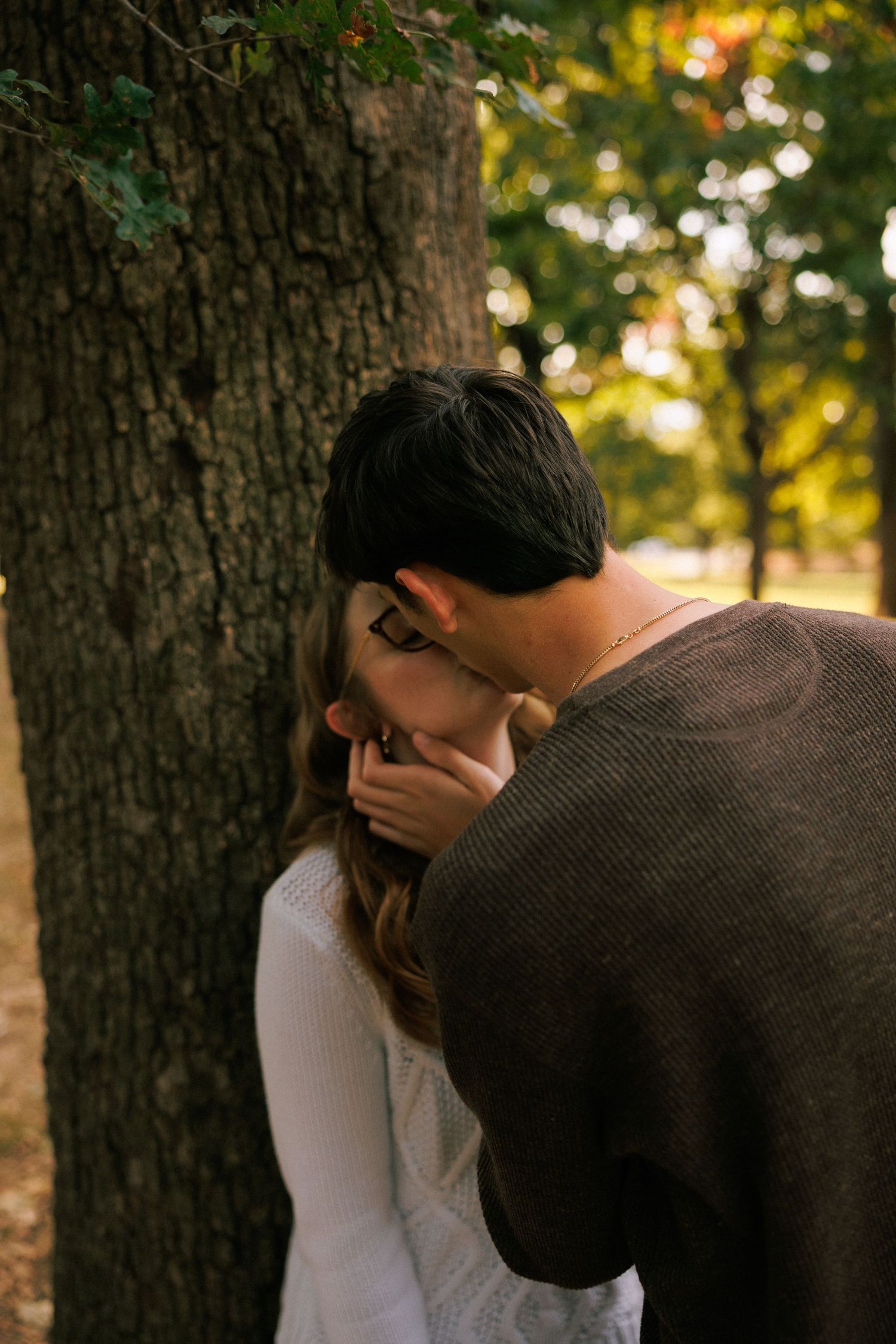 A couple shares an intimate moment together against a tree trunk in warm sunset lighting.