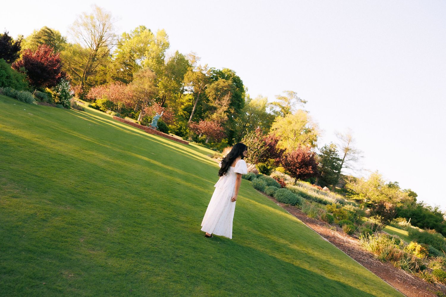 A woman in a white dress stands on a lush green lawn surrounded by colorful gardens at golden hour.