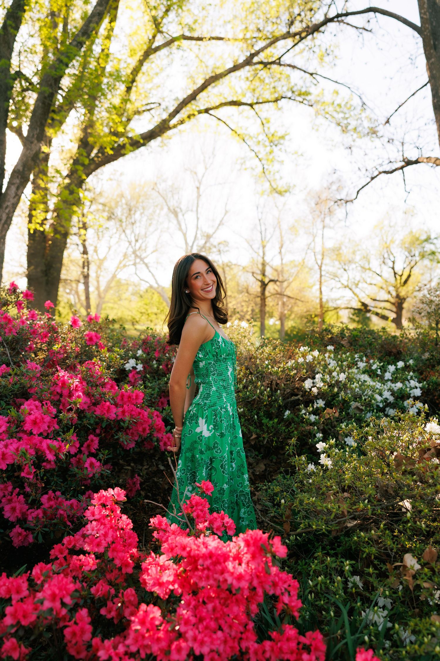 Young woman in green lace dress smiling among vibrant pink and white azalea blooms in a sunlit garden.