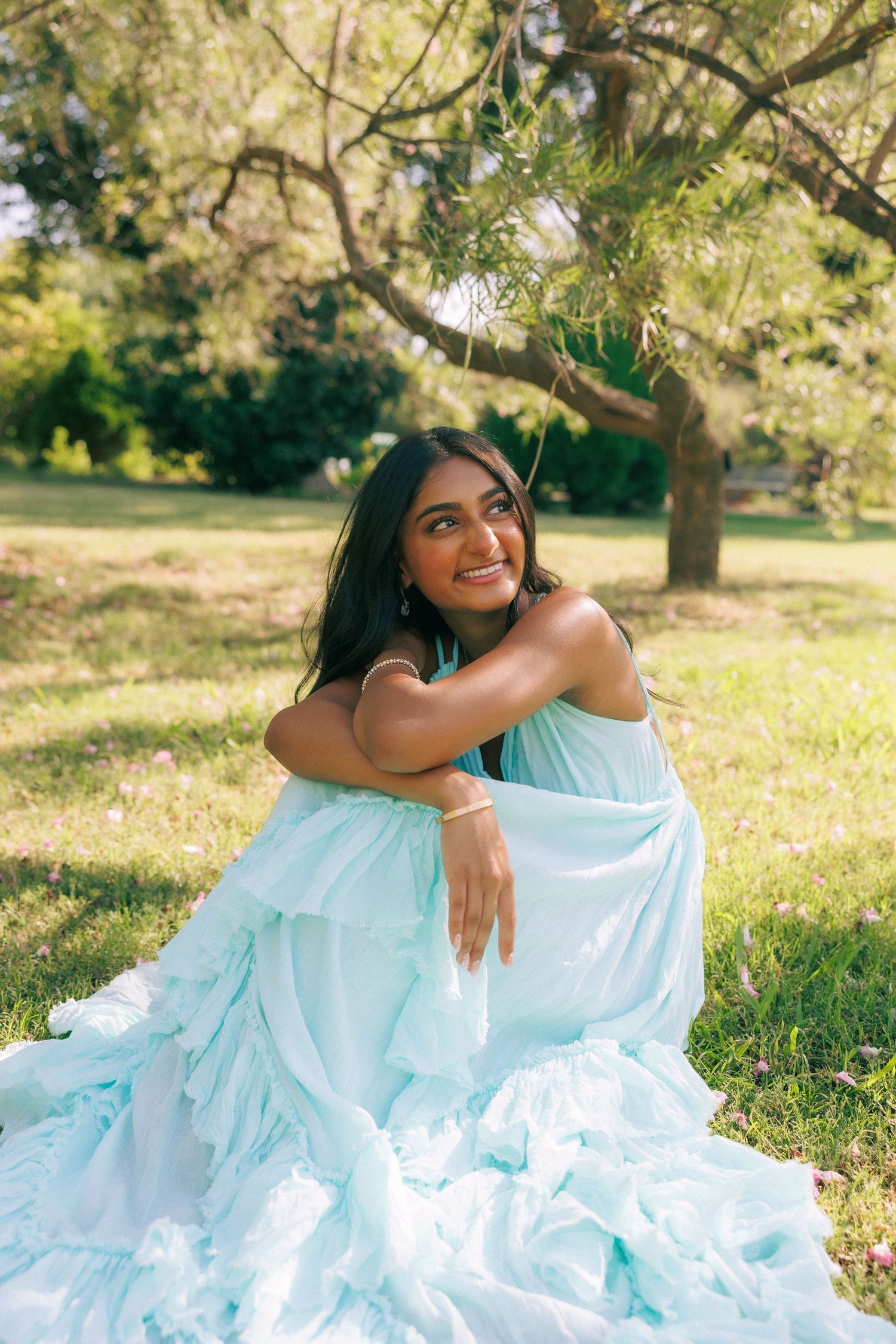Young woman in a light blue ruffled gown sitting gracefully on grass under a tree, smiling outdoors.