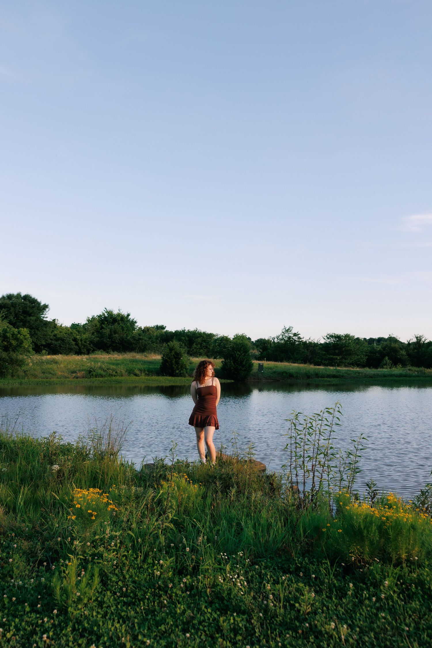 A young woman in a red dress stands by a serene lake surrounded by lush greenery and wildflowers at dusk.