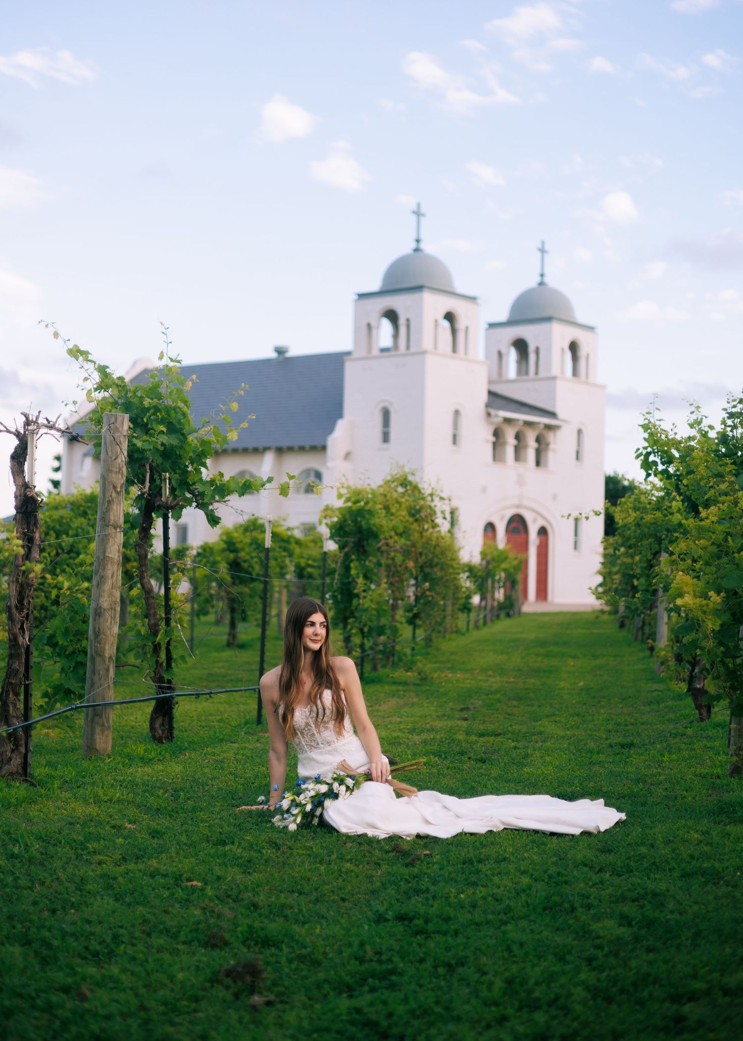 A bride in a white gown sits gracefully on a grassy hillside near a mission-style church.