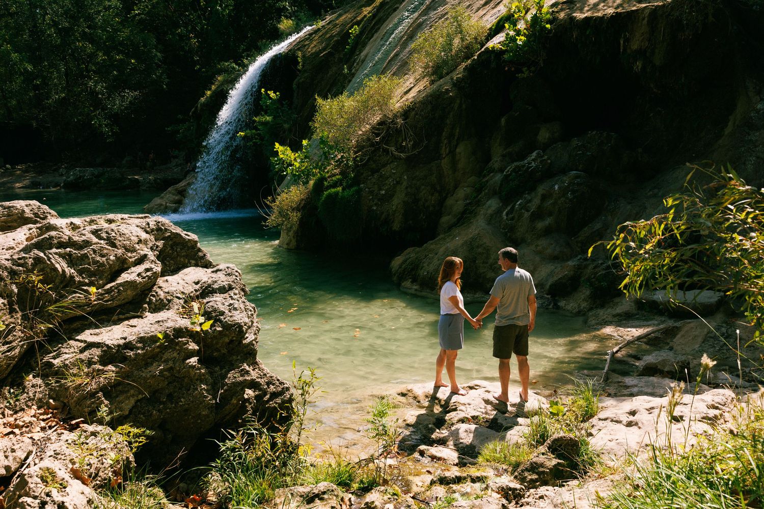 Two people stand on rocks near a scenic waterfall in a hidden natural pool.