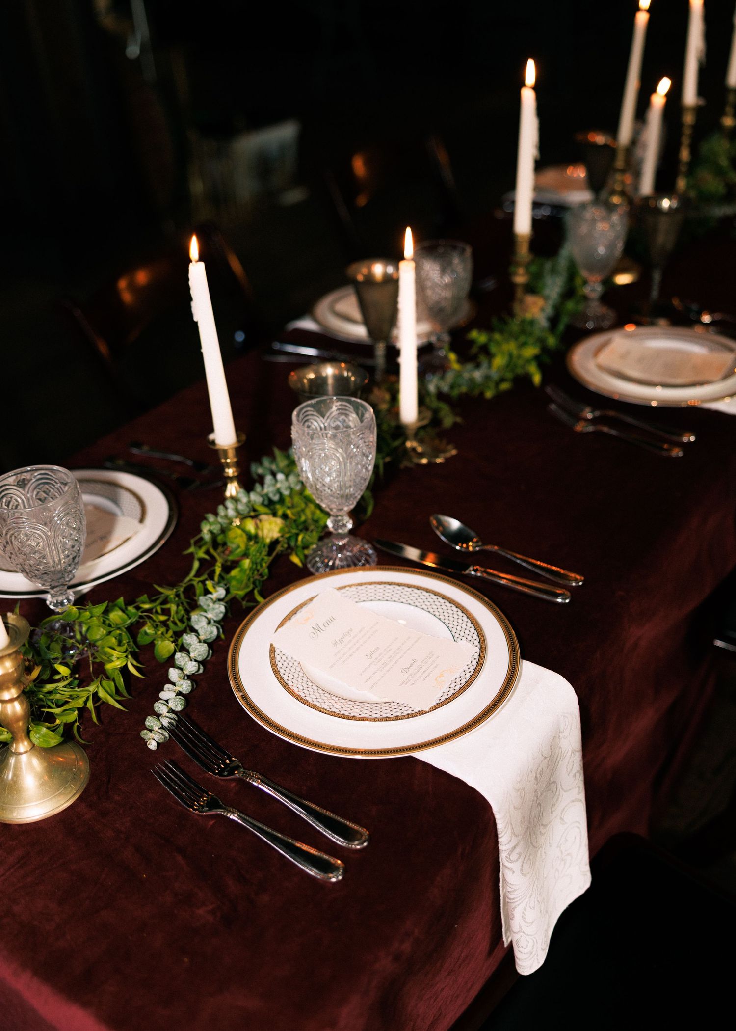 Elegant wedding table settings with lit candles, white plates, and greenery on a dark tablecloth.