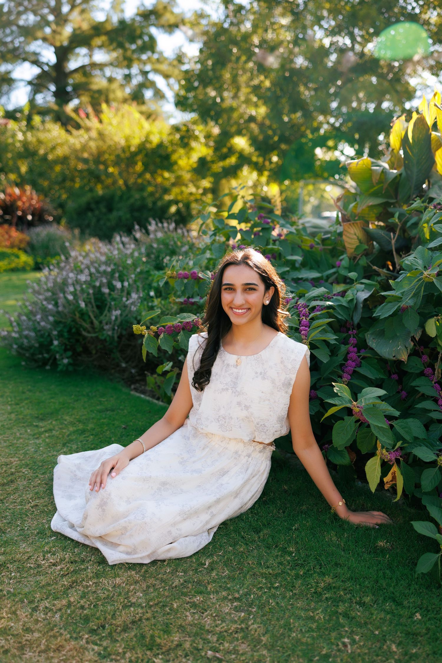 Young woman in floral dress sitting on green grass beside colorful garden flowers, smiling outdoors.