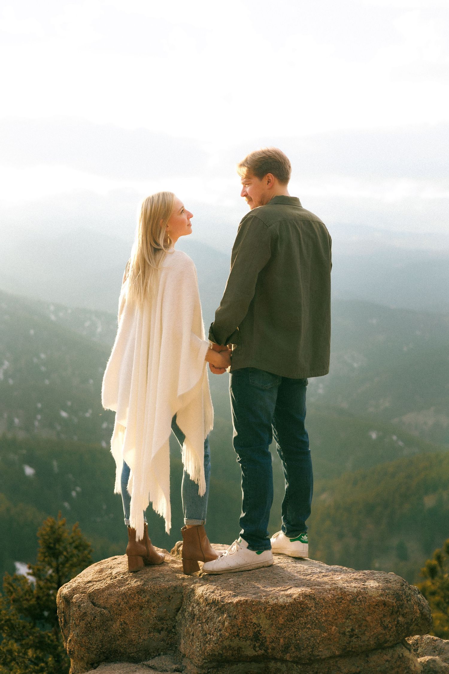 A couple stands together on a rocky mountain overlook with misty peaks in the background.