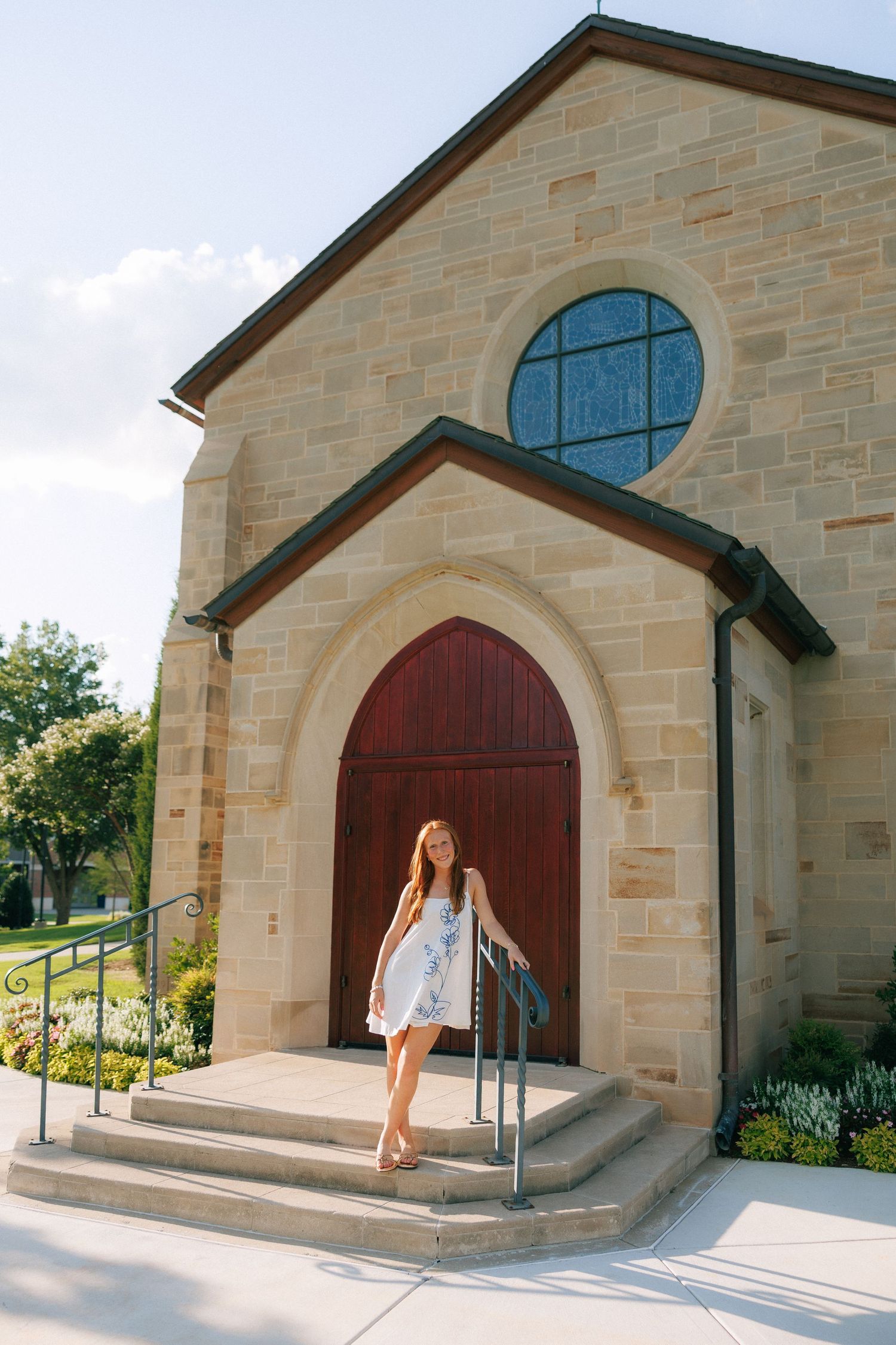 Young woman in white dress smiling on stone church steps with arched red door and round stained glass window.
