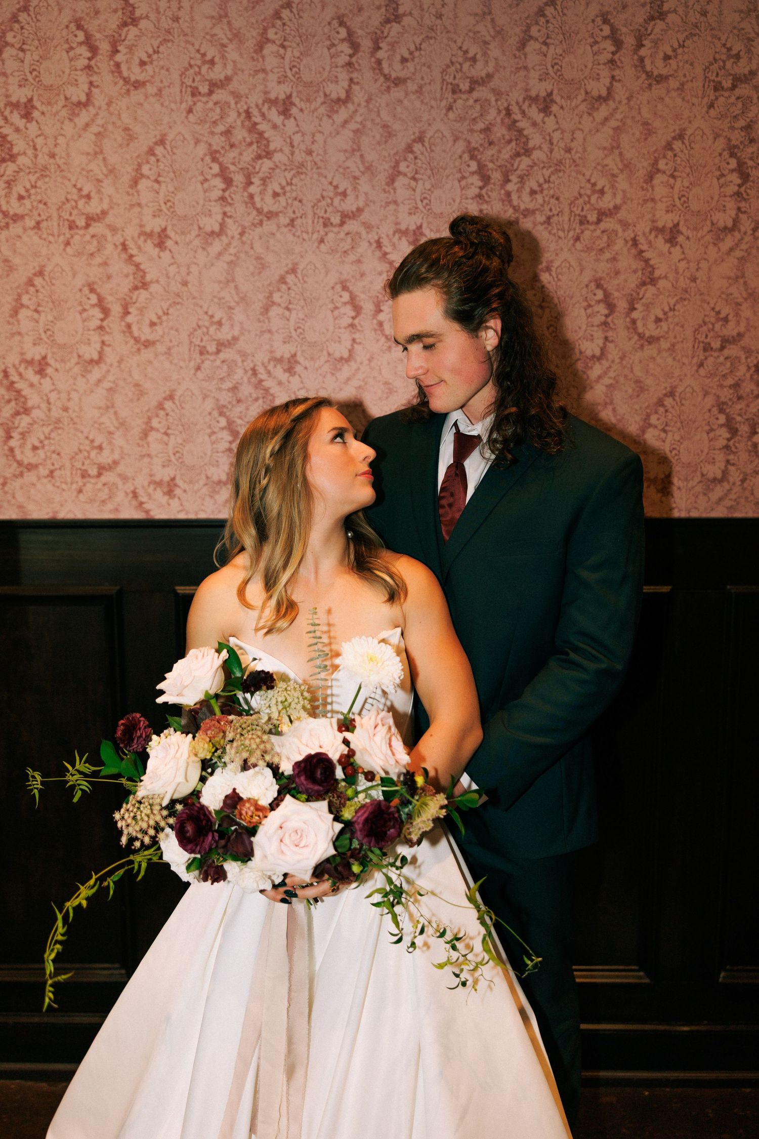 A romantic wedding scene against a vintage wallpapered wall with floral bouquets.