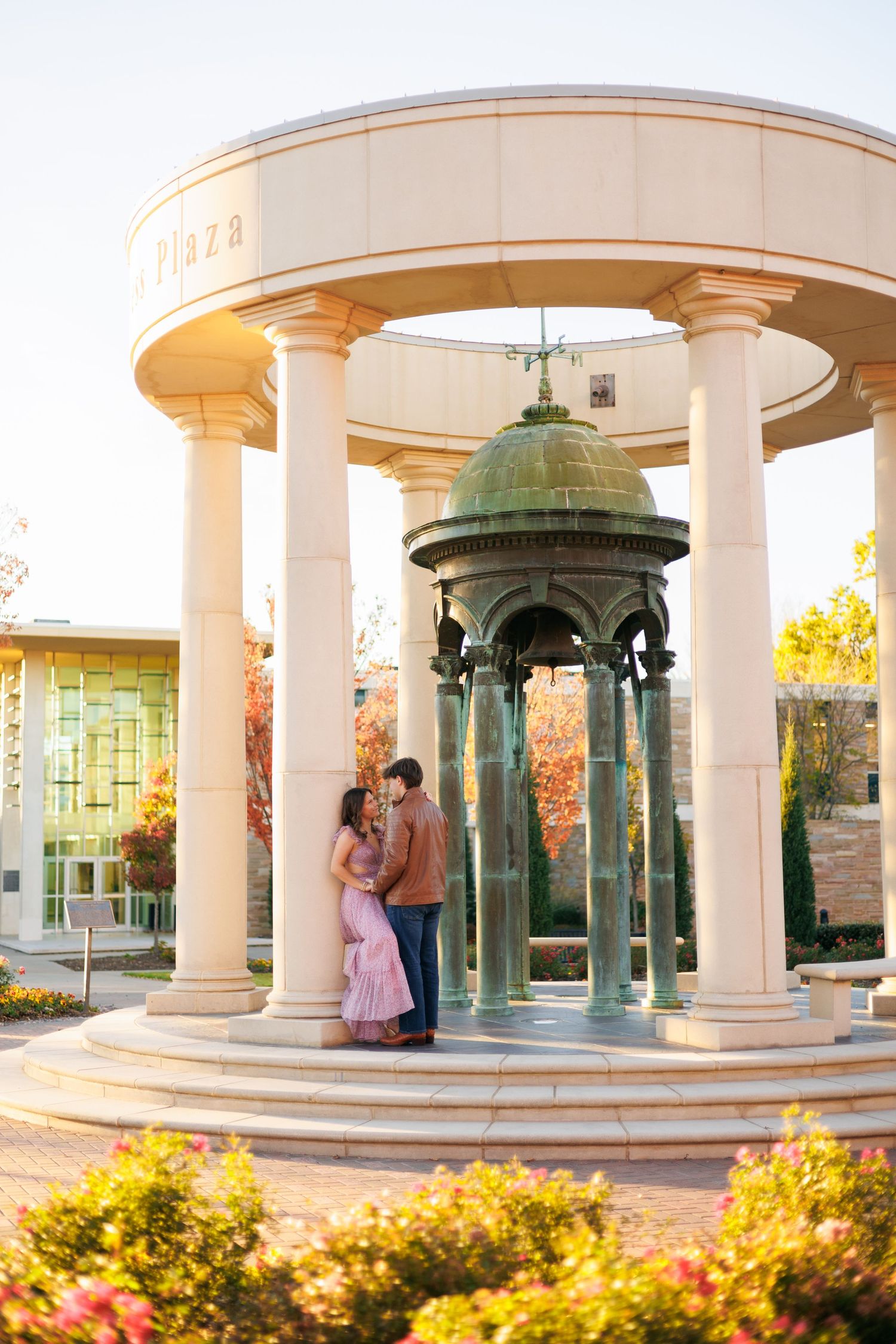 White columned gazebo with green domed roof surrounded by yellow and pink flowers in a garden setting.