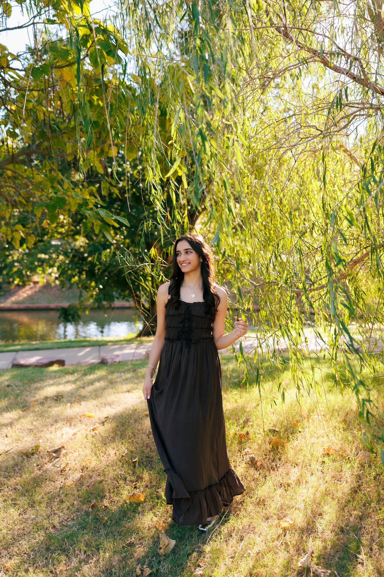 A long black dress contrasts beautifully with sunlit willow trees in a park setting.