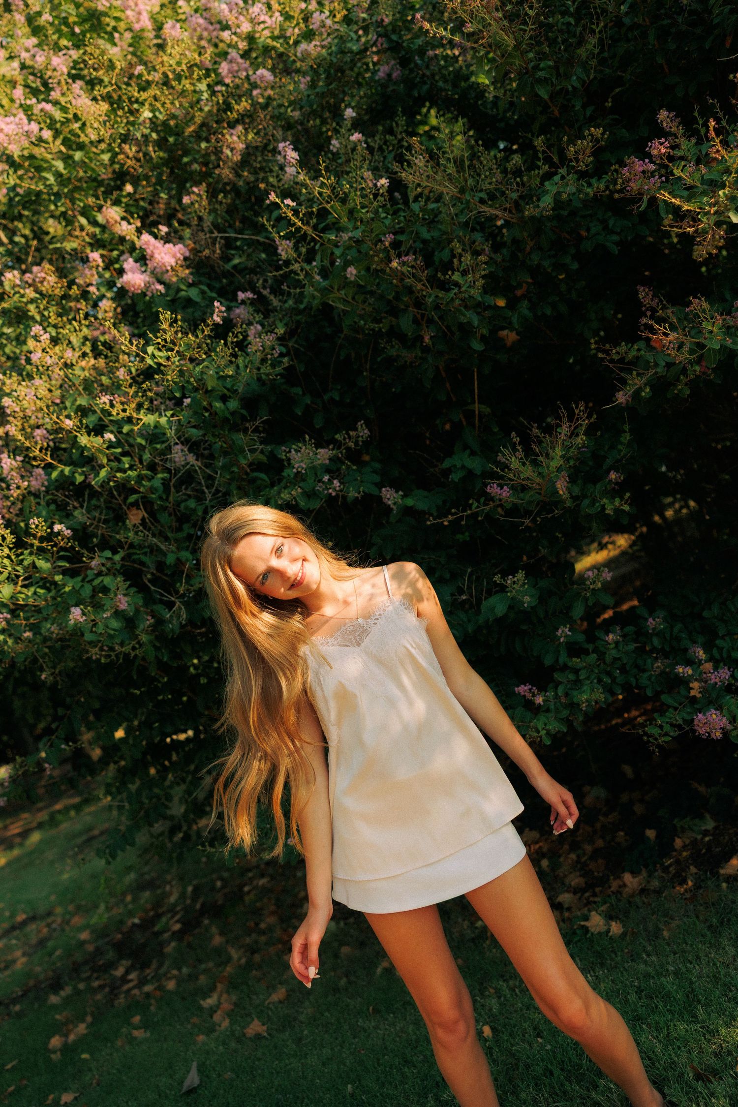 Blonde woman in white mini dress posing outdoors near blooming pink flowers and lush green bushes in golden sunlight.