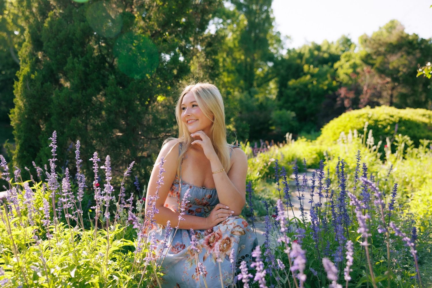 Blonde woman in floral dress sitting among purple lavender flowers in a lush green garden on a sunny day.