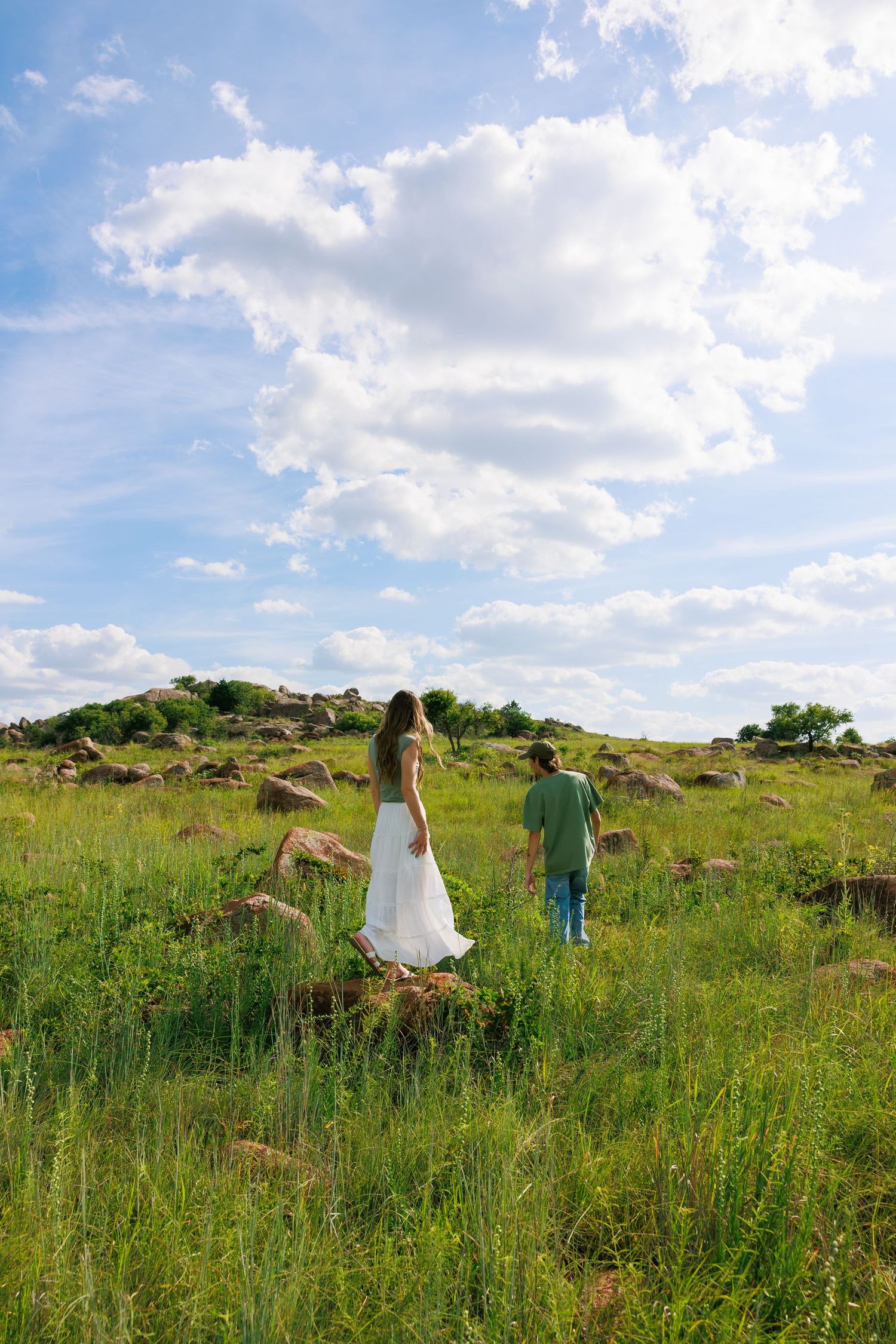 Two people walk through a lush green meadow with rolling hills in the background.