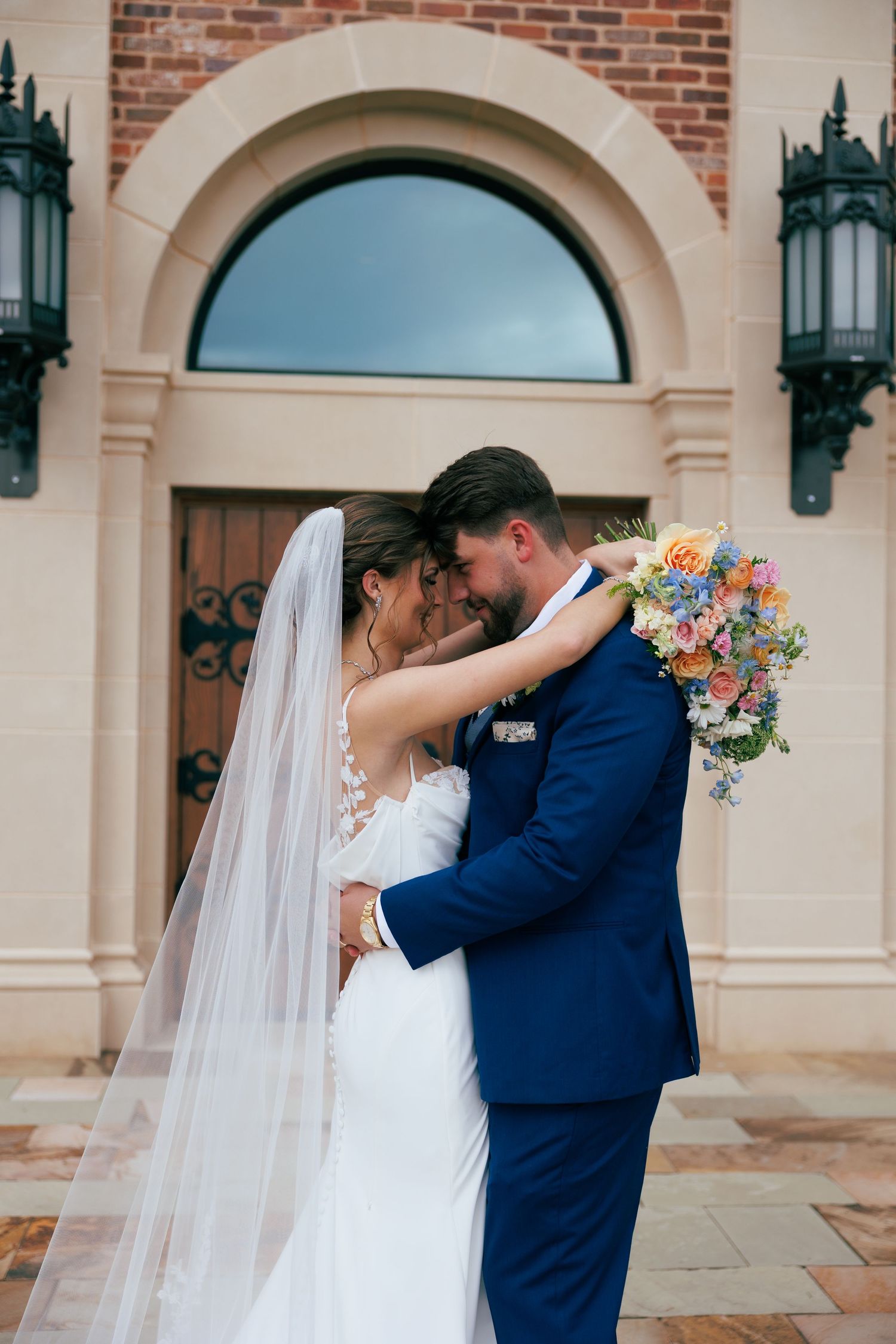 Romantic wedding couple embracing and kissing outside a brick building with arched doorways.