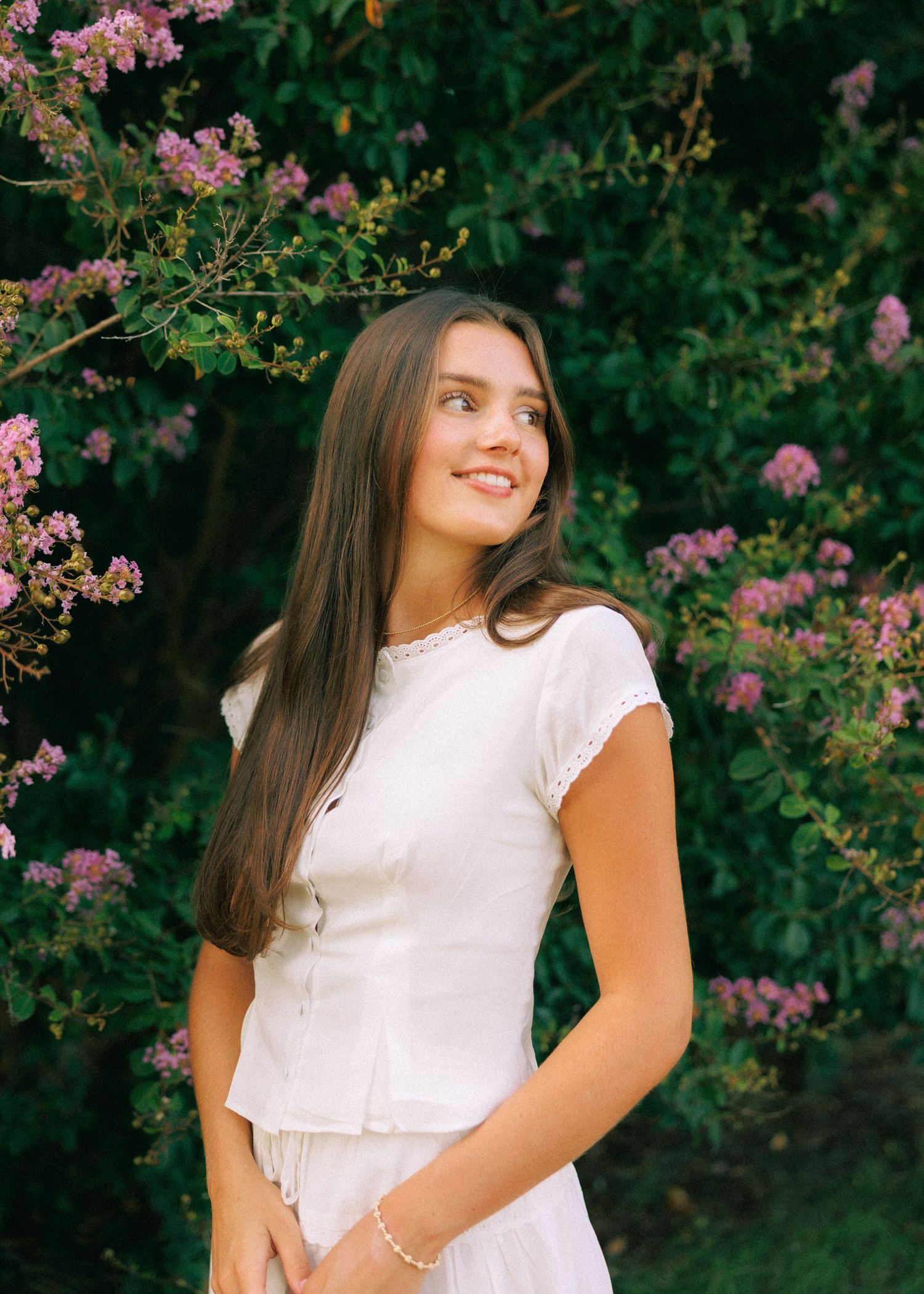 Young woman with long brown hair smiles in white outfit among blooming pink flowers and lush green trees.