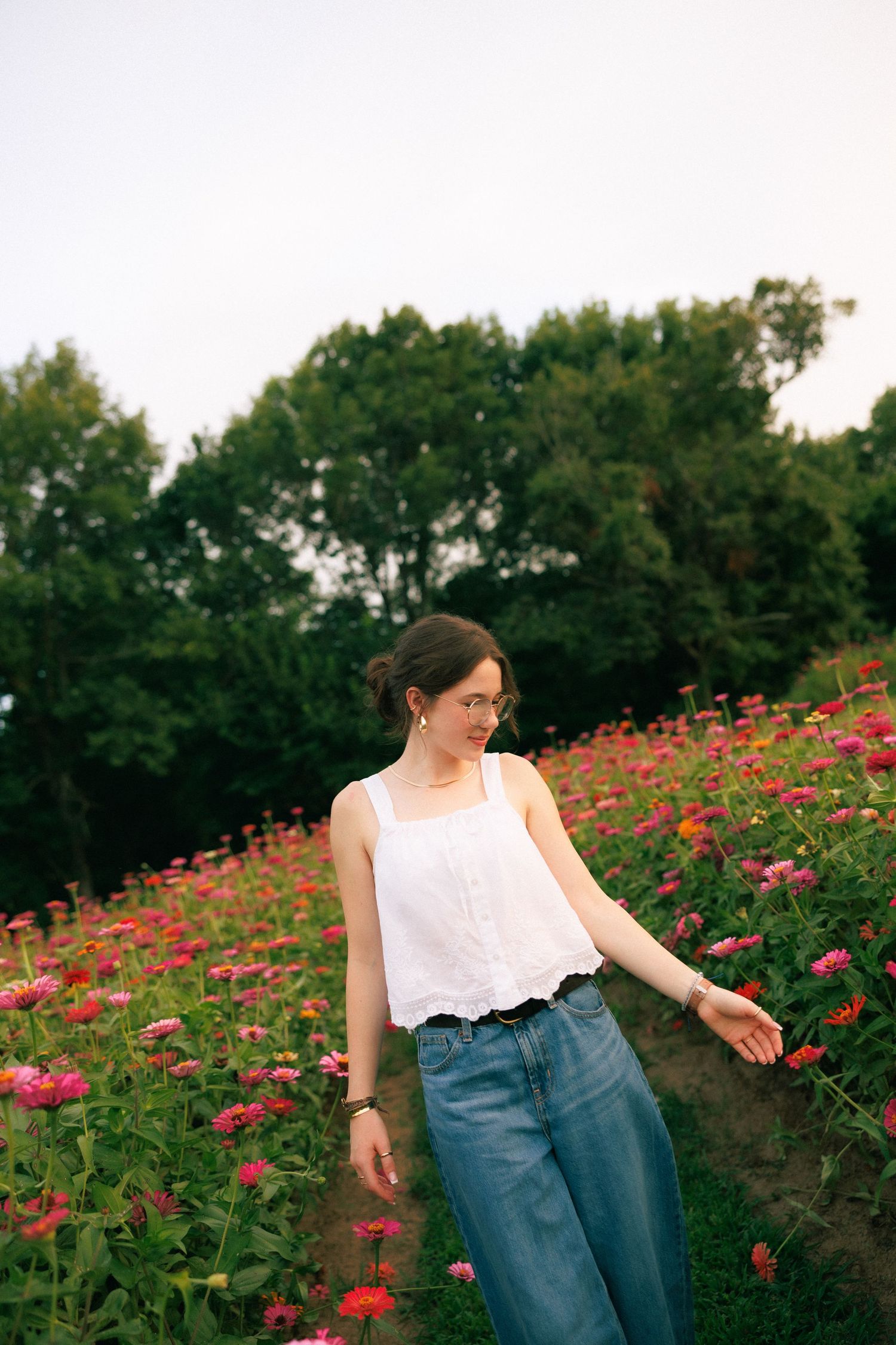 Young woman in white tank top and jeans standing in a vibrant flower field with pink and red blooms and green trees.