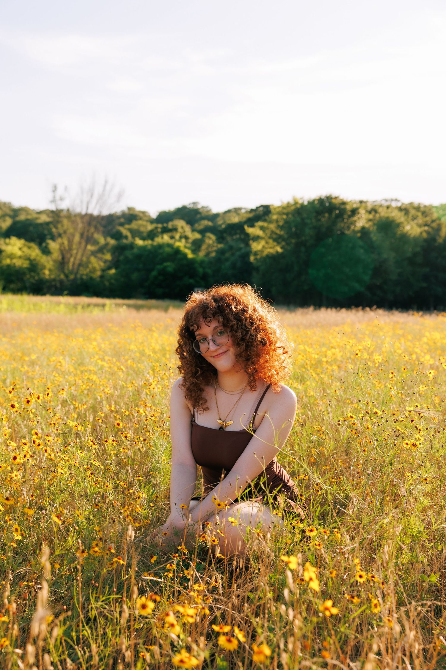 Young woman with curly red hair sitting in a sunny wildflower meadow, wearing a brown dress and glasses.
