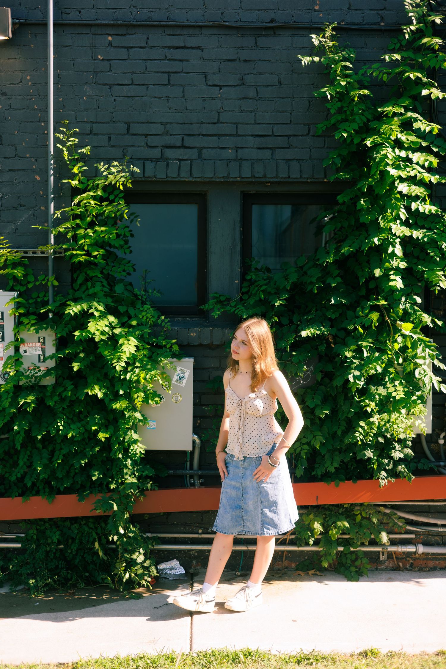 Someone in casual summer attire poses against a dark brick wall with climbing green vines.