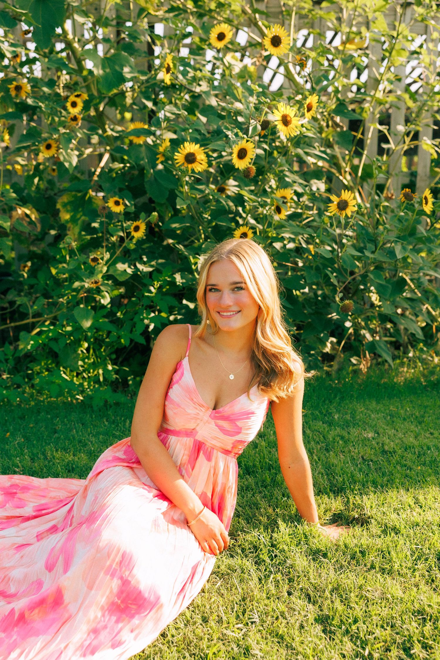 Young blonde woman in pink floral maxi dress sitting on green grass in front of blooming sunflowers.